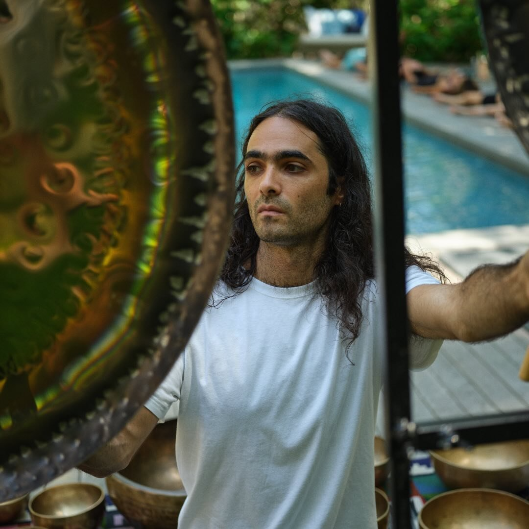 A man with long dark hair and light skin, wearing a white shirt, standing outdoors near a pool, looking serious and focused.