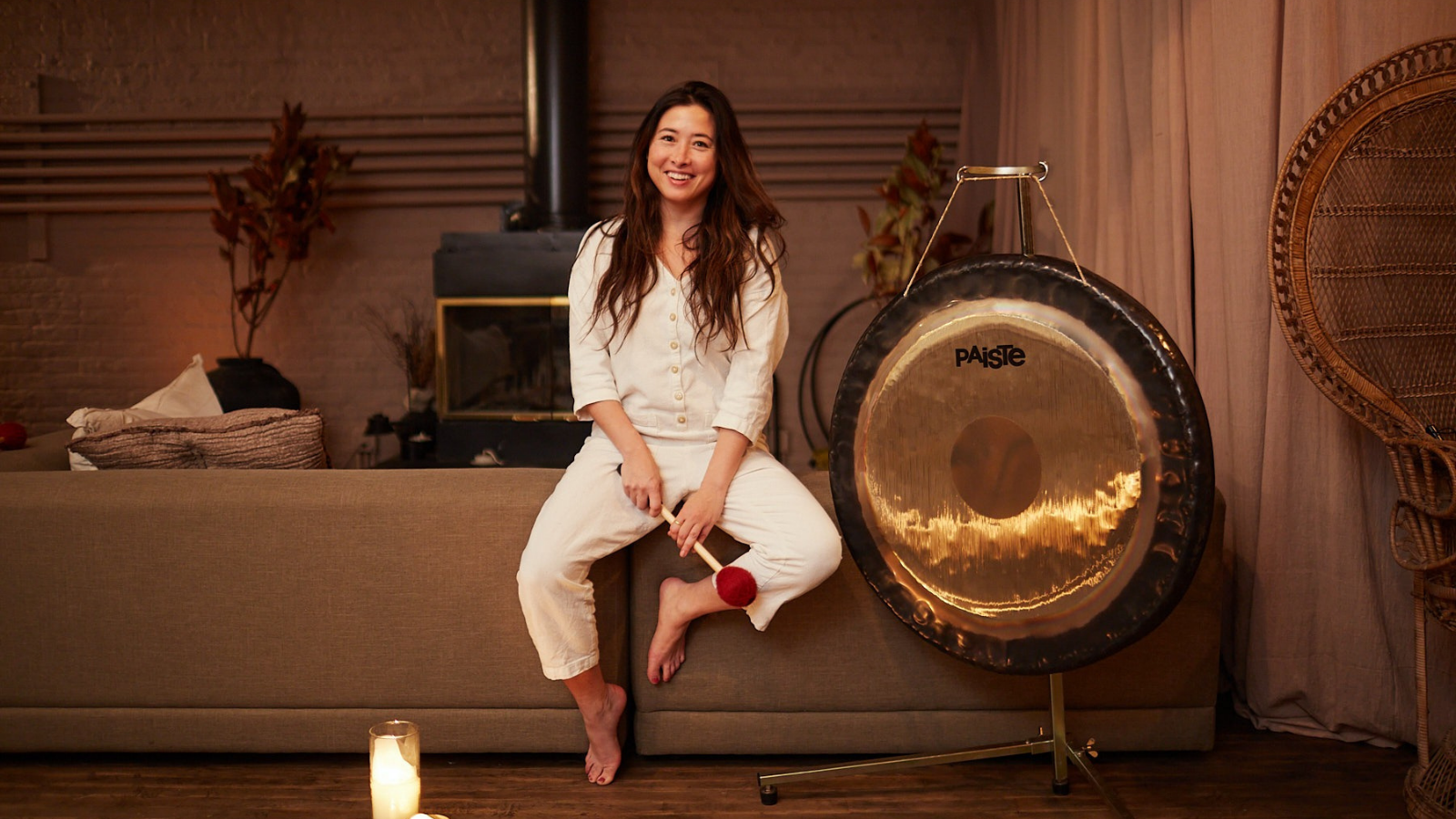 A woman with long brown hair in white pajamas sits on a beige sofa, holding a percussion mallet and smiling in a cozy living room with a large gong nearby.