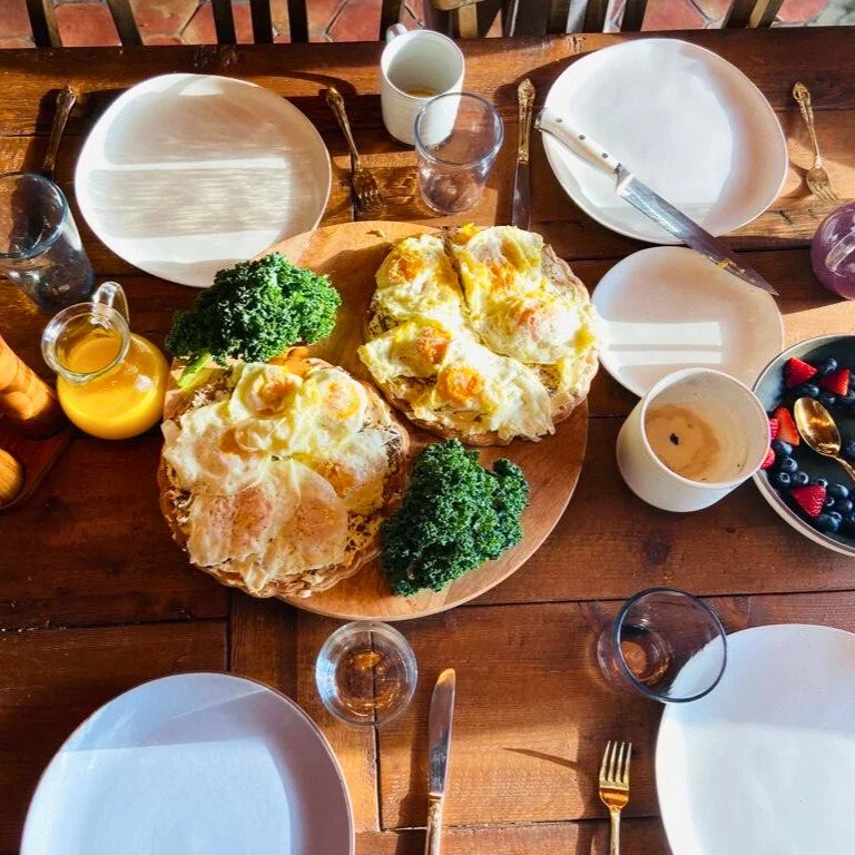 Breakfast table with two egg dishes topped with cheese and vegetables, surrounded by empty plates, glasses, a bowl of berries, juice, coffee, and utensils.