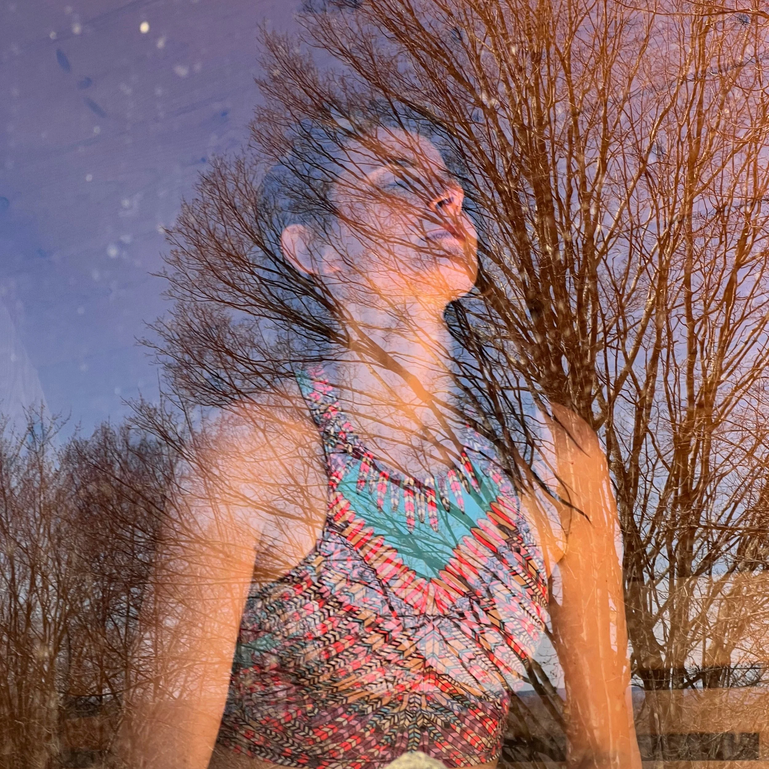 Reflection of a woman with glasses and curly hair, standing next to leafless trees against a partly cloudy sky, with a colorful scarf around her neck.