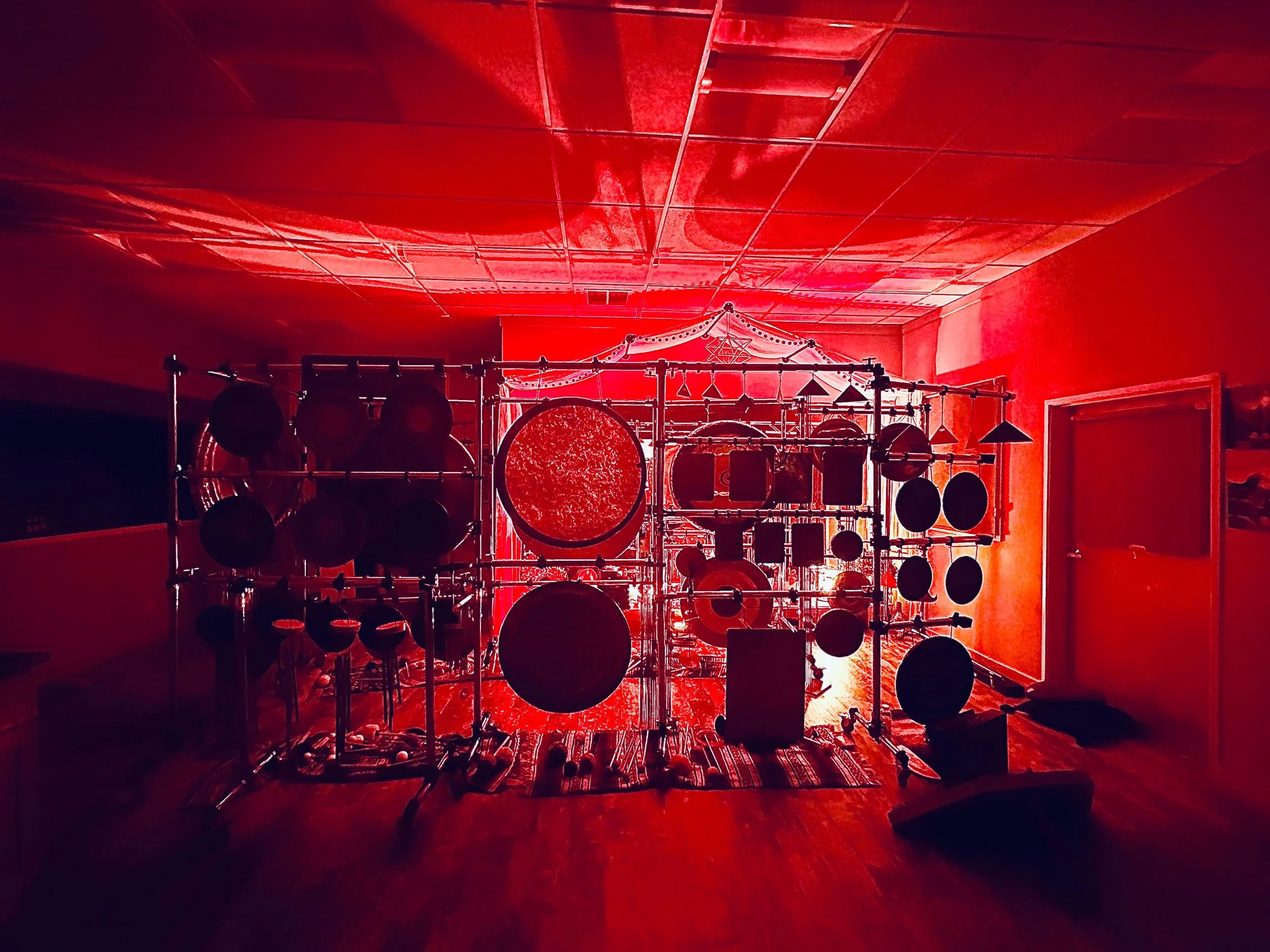A room illuminated with red lighting, containing various percussion instruments such as drums and cymbals mounted on racks, with a decorated wall overhead.
