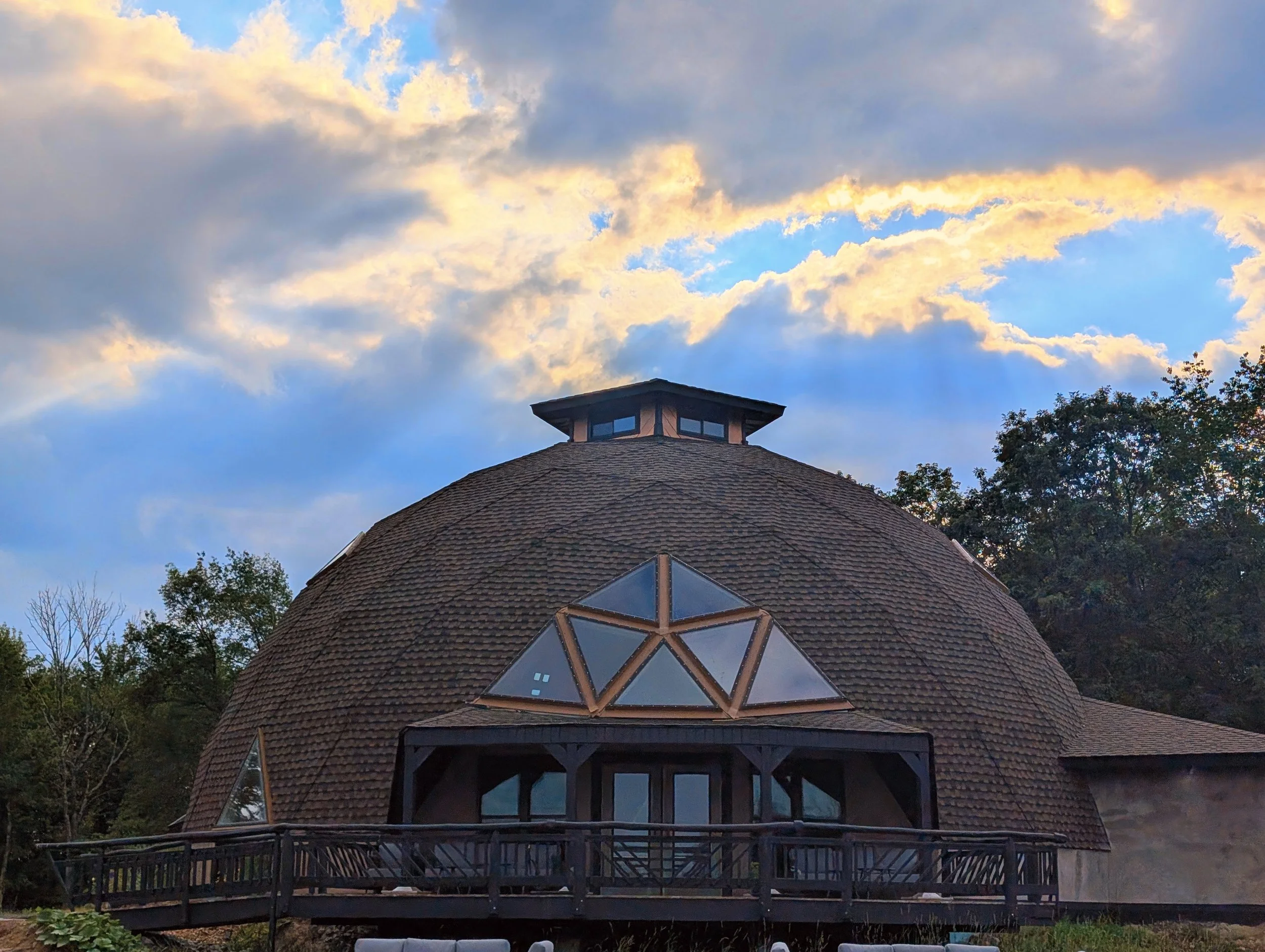 View of a house with a round roof and a small tower at the top, featuring windows, surrounded by trees and a partly cloudy sky with sunlight breaking through.
