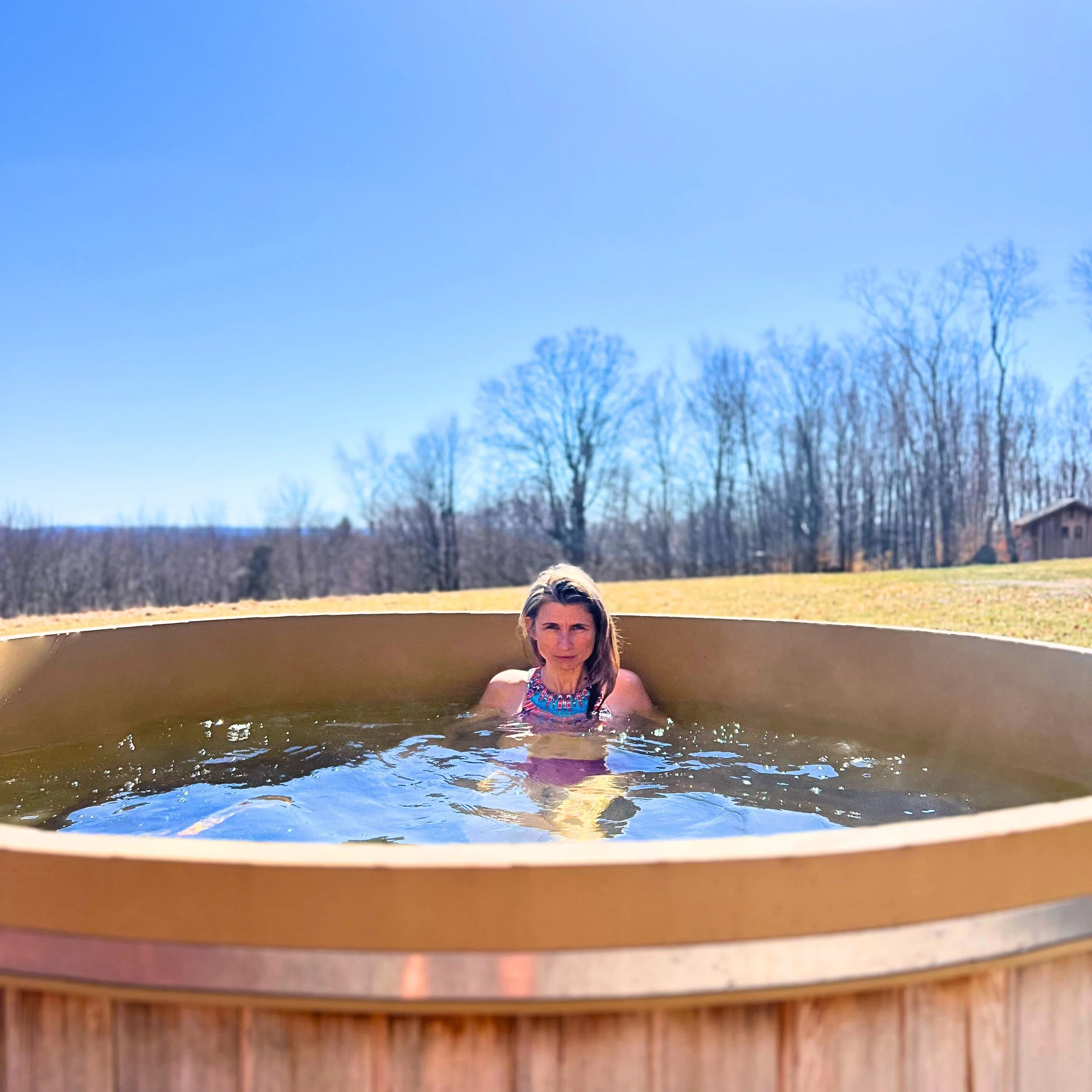 A woman is sitting in a hot tub outdoors with a clear blue sky and leafless trees in the background.