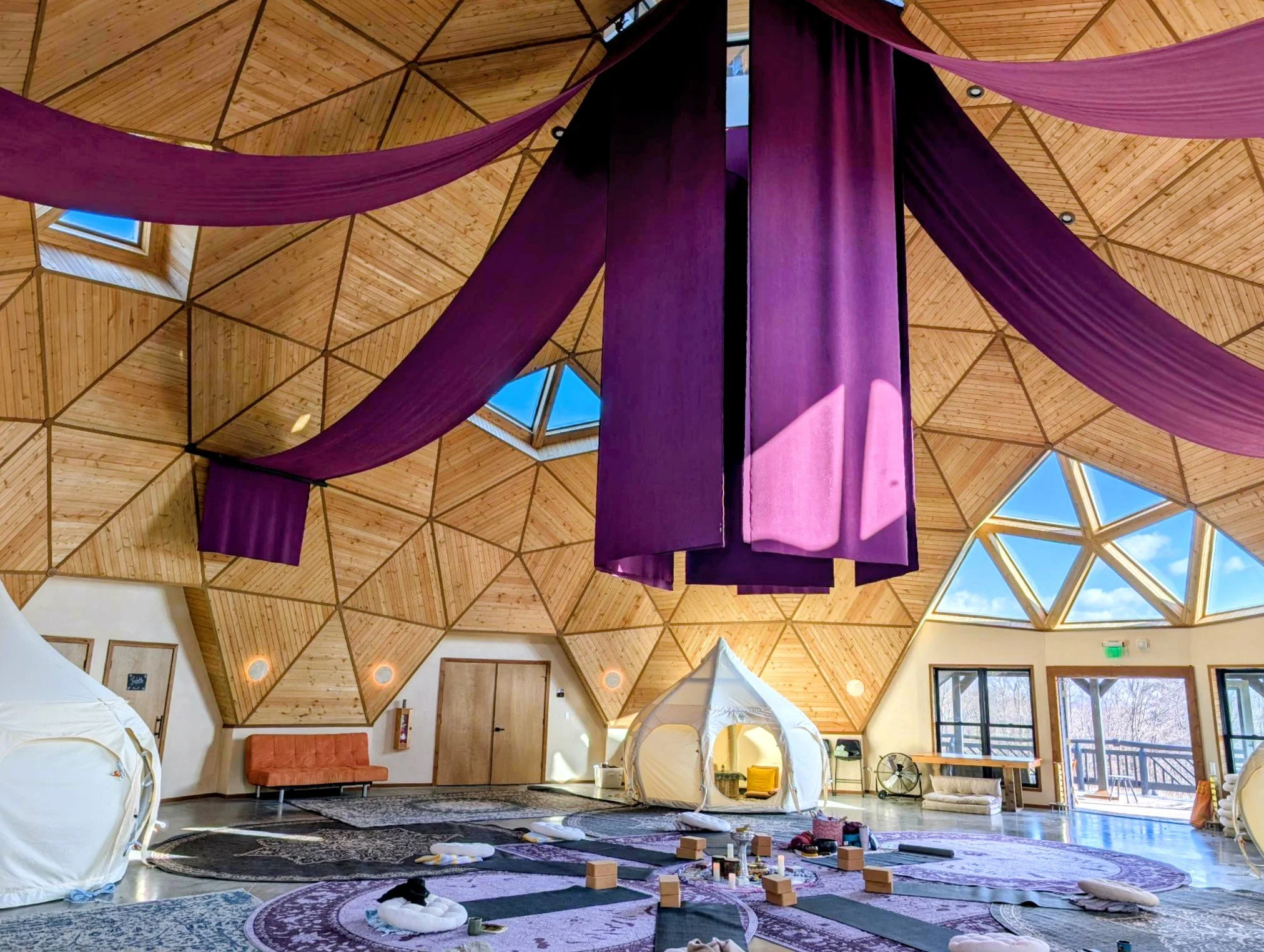 Interior of a wooden dome structure with purple fabric drapes hanging from the ceiling, windows letting in natural light, and seating with cushions and children playing with building blocks on a rug.