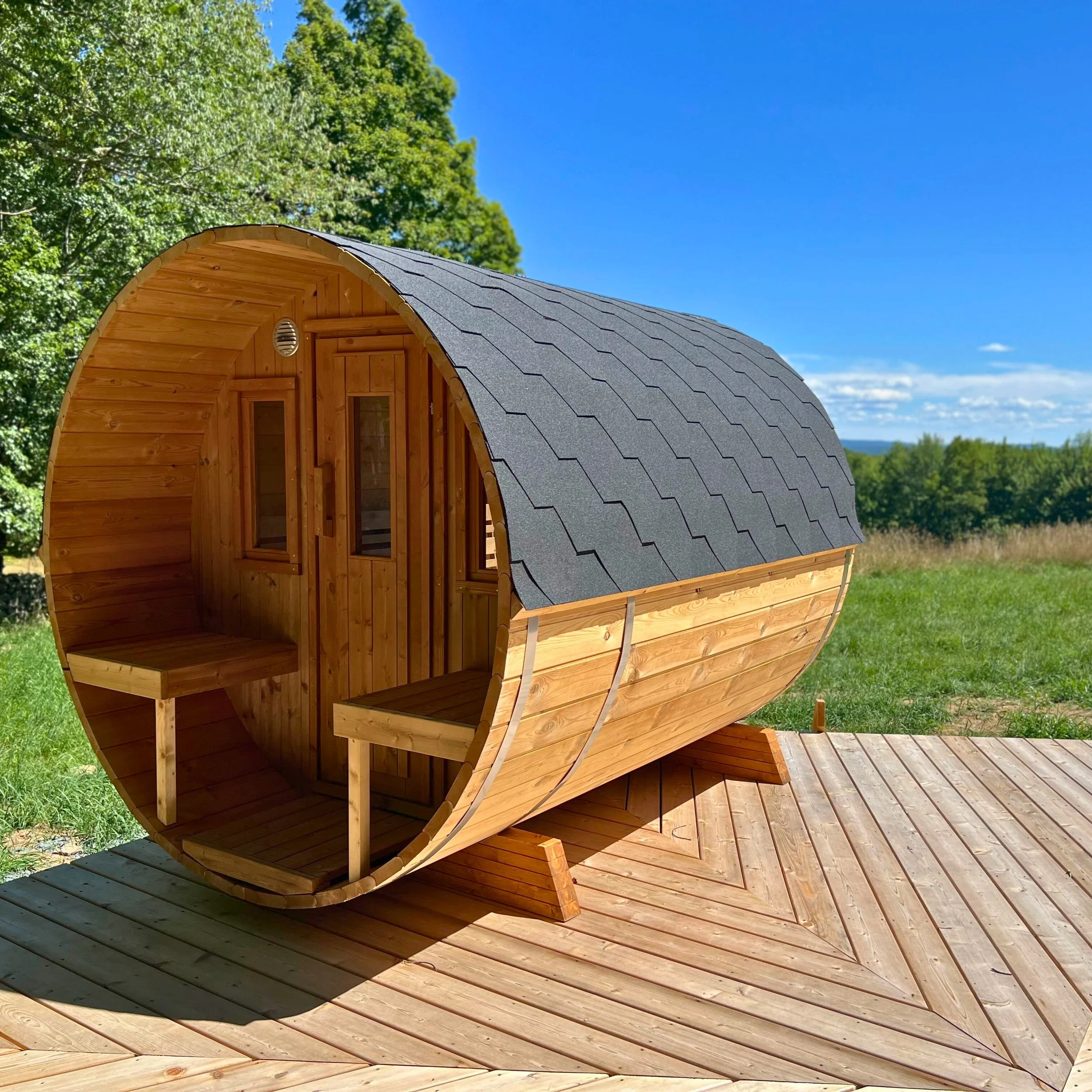 A small wooden sauna with a curved roof and dark shingles, located outdoors on a wooden deck, with trees and a bright blue sky in the background.