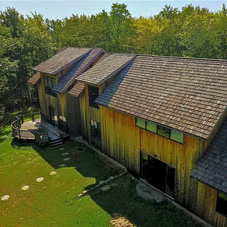 Aerial view of a wooden house with a sloped roof and multiple dormer windows, surrounded by a green lawn and trees.