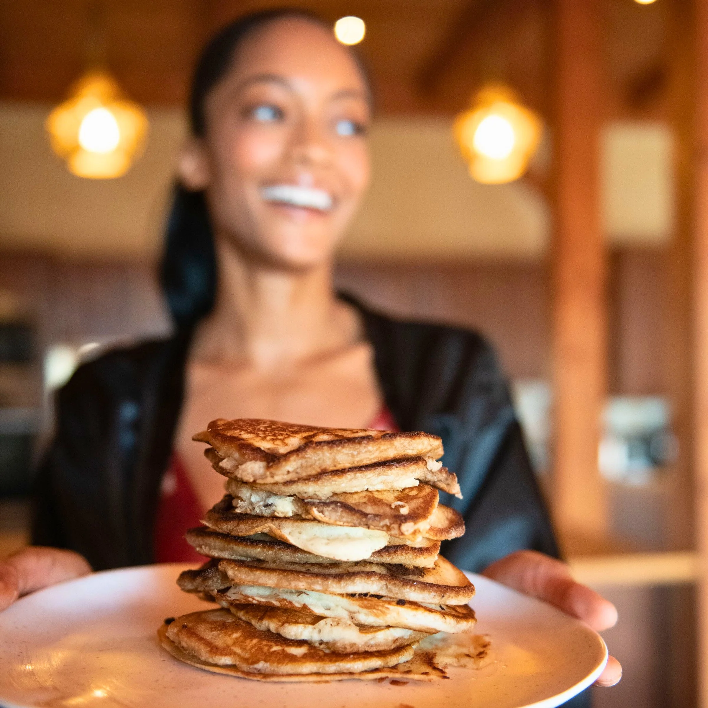 Close-up of a stack of pancakes on a white plate with blurred background of a woman with long dark hair smiling.