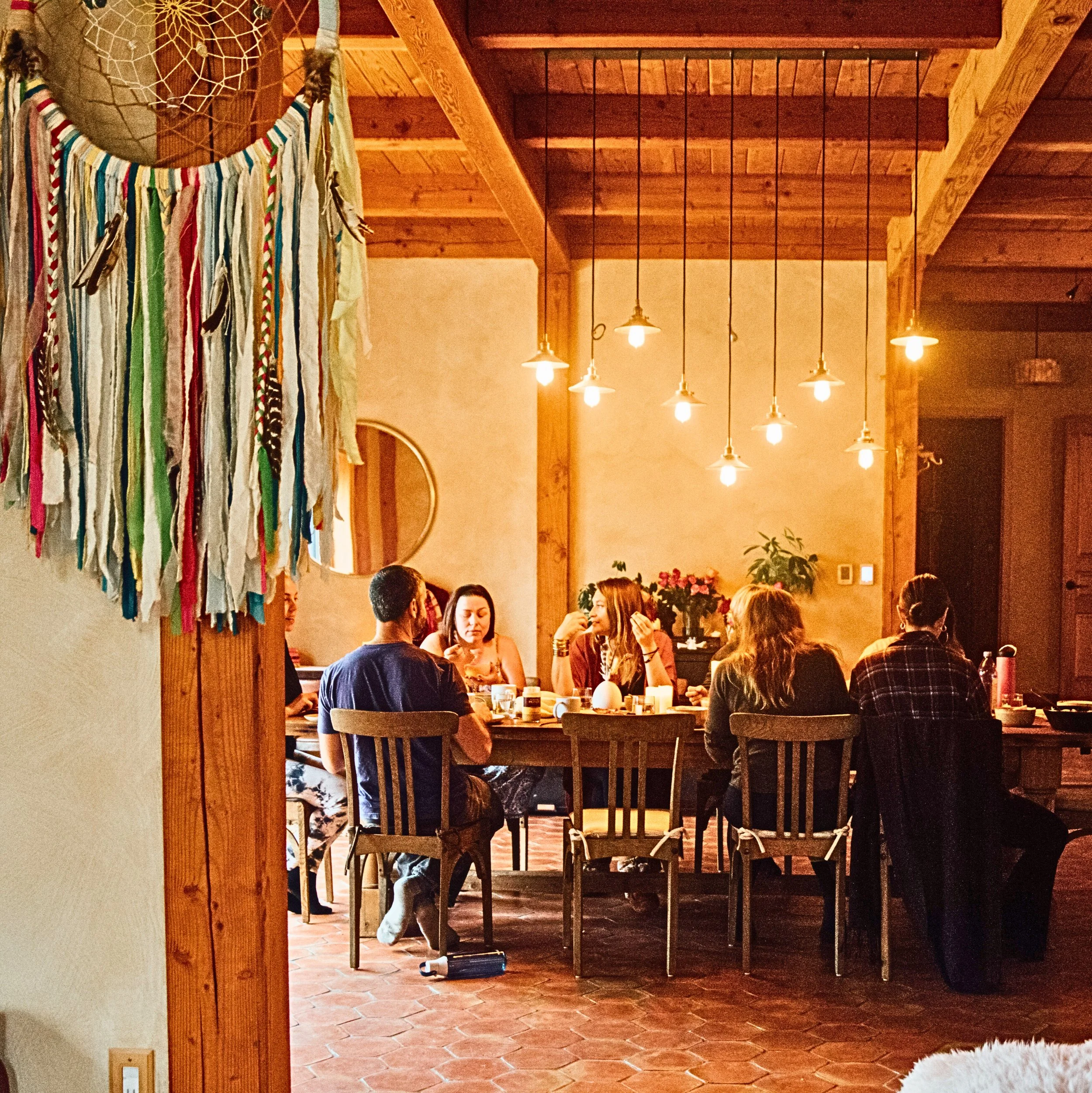 A cozy indoor gathering with people sitting at a table under warm hanging lights, wooden ceiling, and beams, with colorful fabric or ribbons hanging on the left wall.