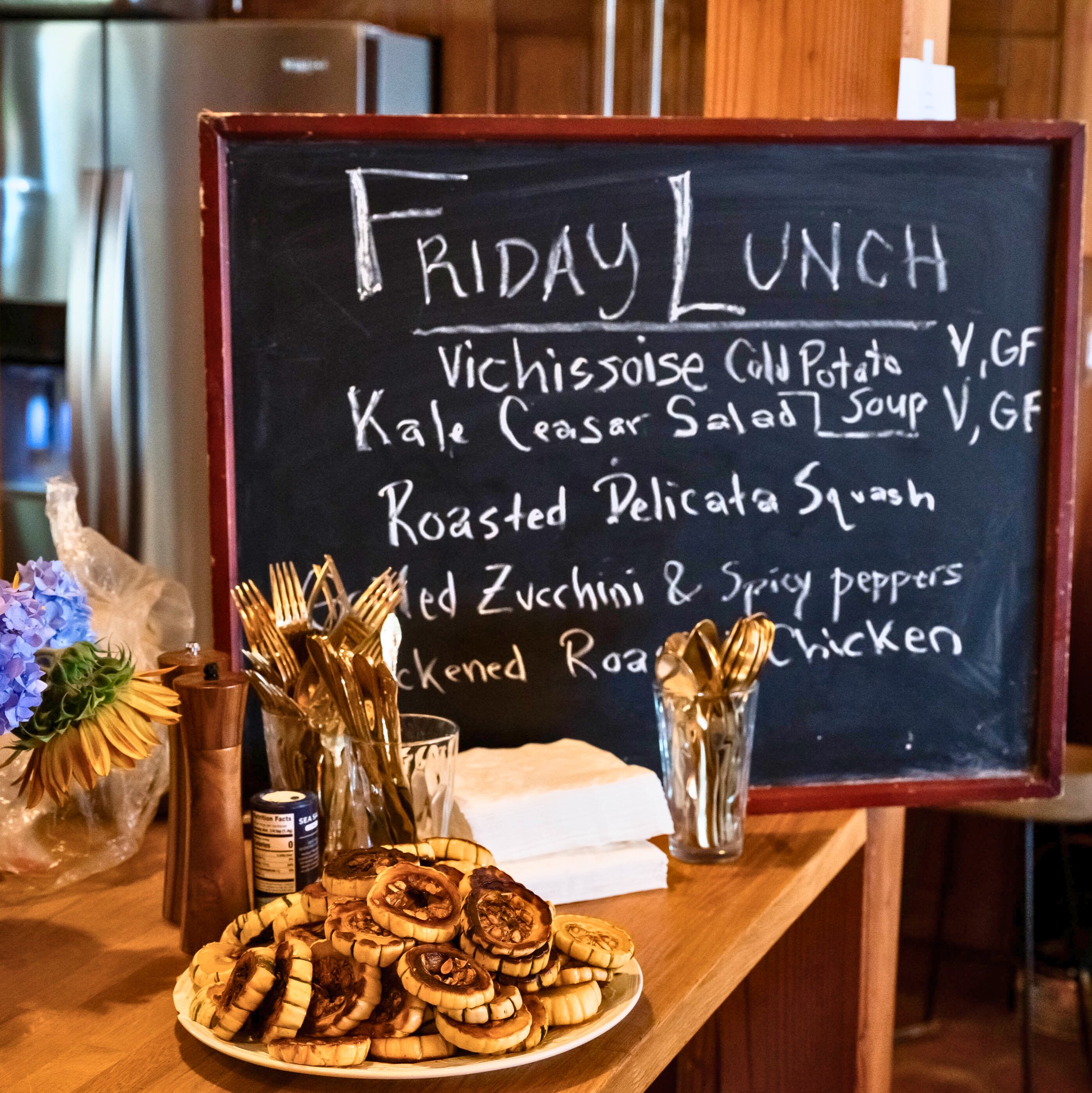 A blackboard sign advertising Friday lunch with menu items including vichyssoise cold potato soup, kale Caesar salad, roasted delicata squash, and grilled chicken, with decorative flowers and pastries in the foreground.