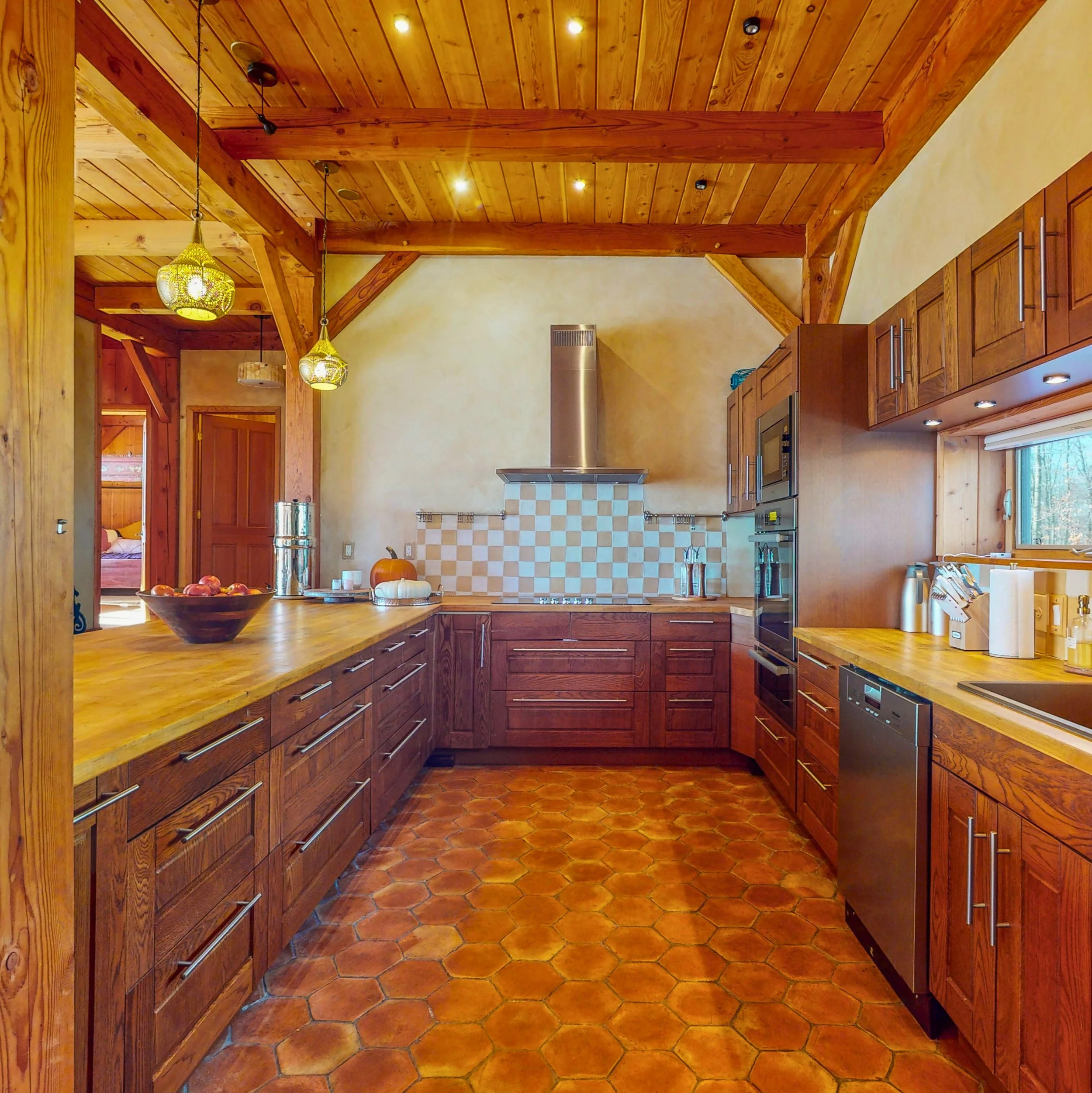 Kitchen with wooden cabinets, wooden ceiling with recessed lights, hanging pendant lights, and terracotta tile flooring.