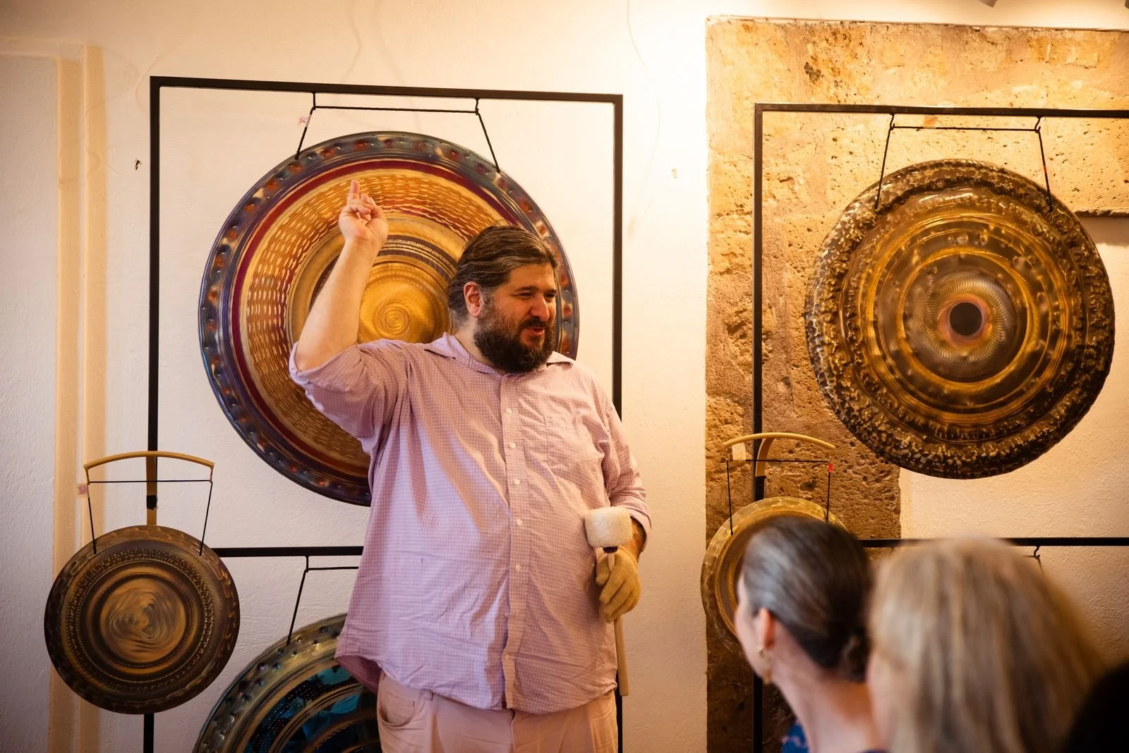 A man with dark hair and beard in a pink shirt, holding a mallet, standing in front of a wall with several large brass gongs on display. The man appears to be explaining or demonstrating something to a group of people.