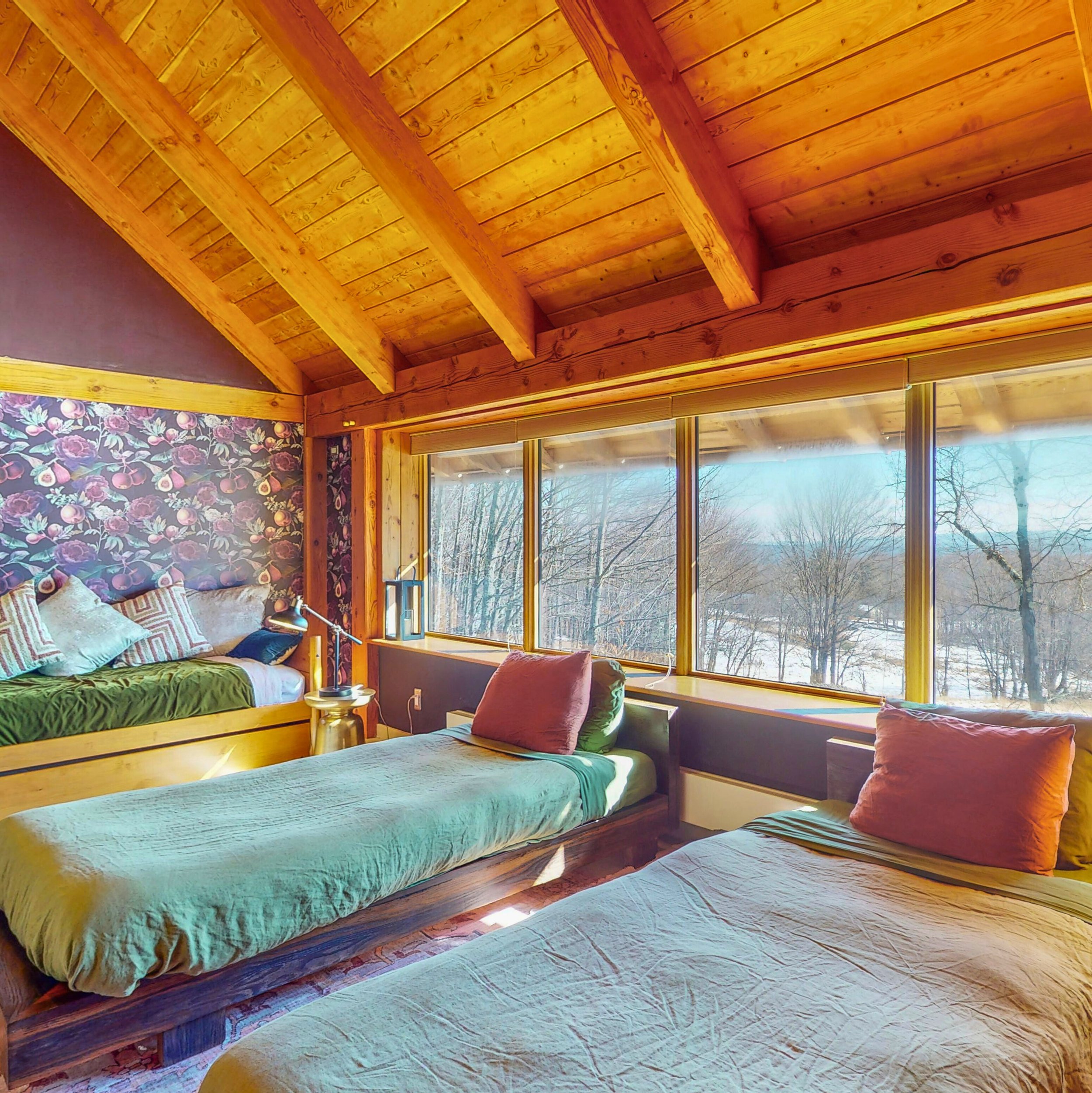 A bedroom featuring a wooden vaulted ceiling with exposed beams, large windows allowing natural light, floral wallpaper, and a bed with turquoise bedding and various pillows.