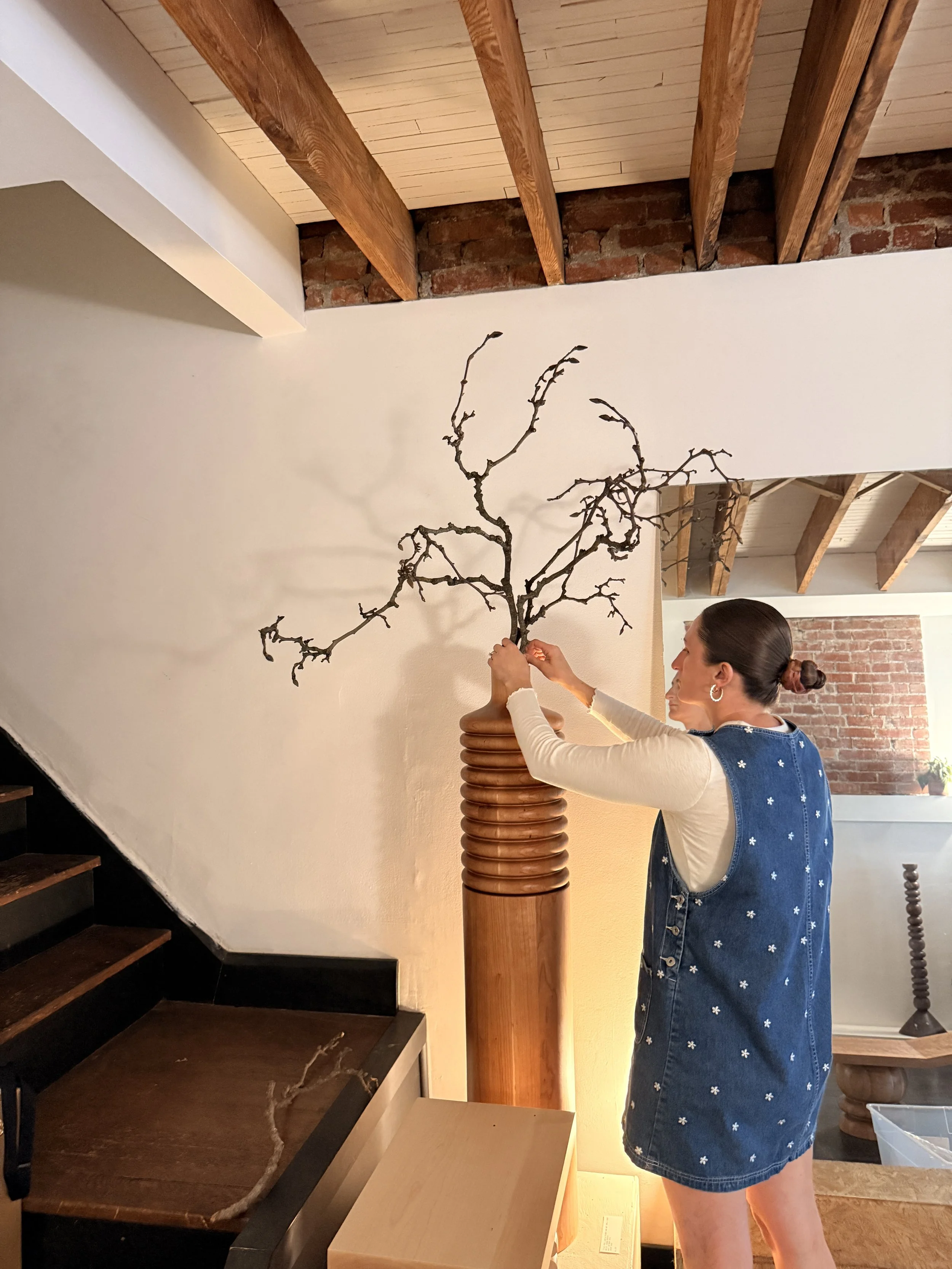 A woman with dark hair tied back, wearing a denim vest and hoop earrings, is holding a leafless tree branch with twisting limbs in a wooden vase. She stands inside a room with visible brick walls, wooden beams on the ceiling, a staircase with dark wood steps, and a beige wall.