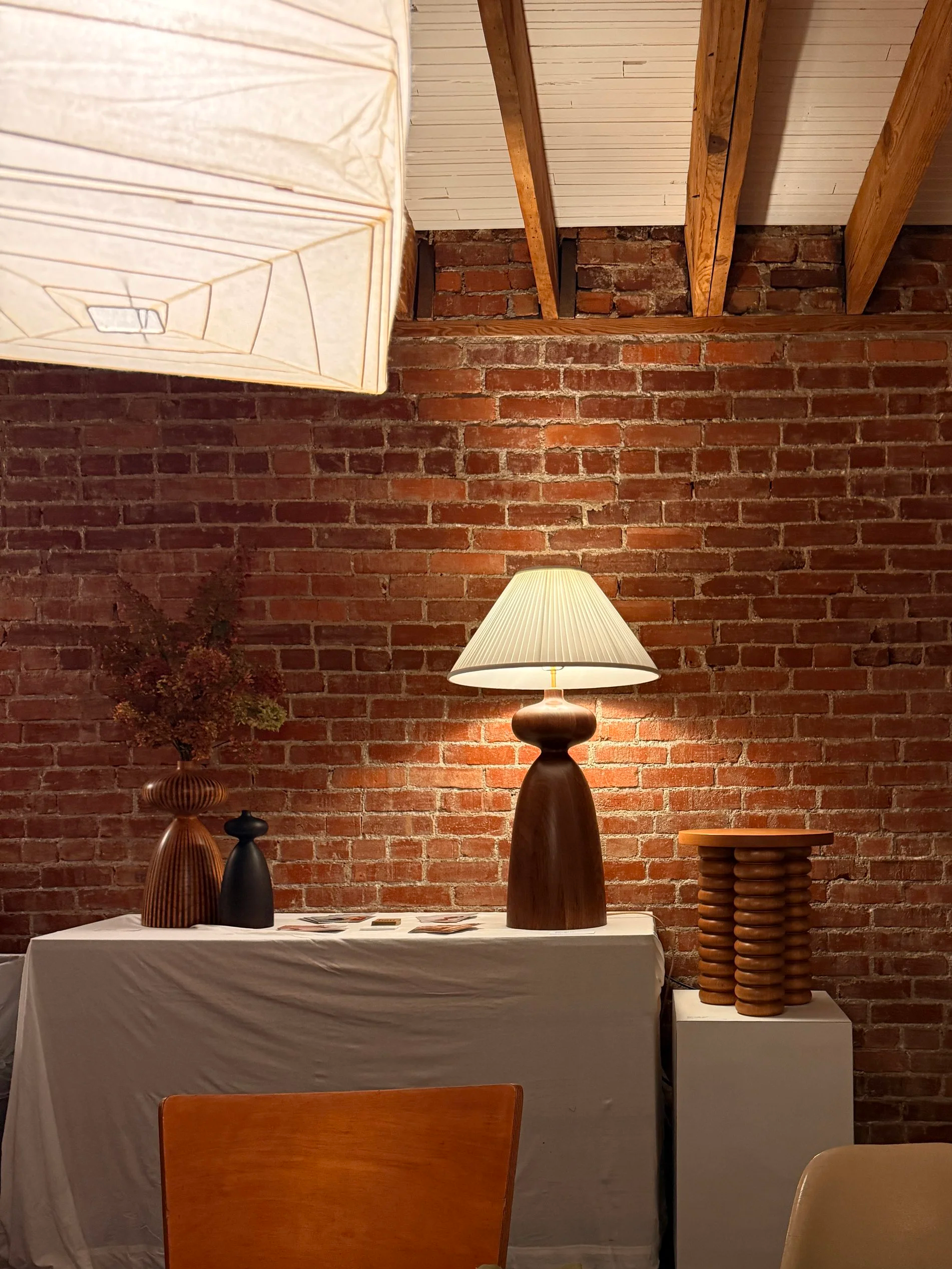 Interior scene with a brick wall, table covered with a white cloth, decorative vases, and a lamp. The ceiling has exposed wooden beams and white paneling.