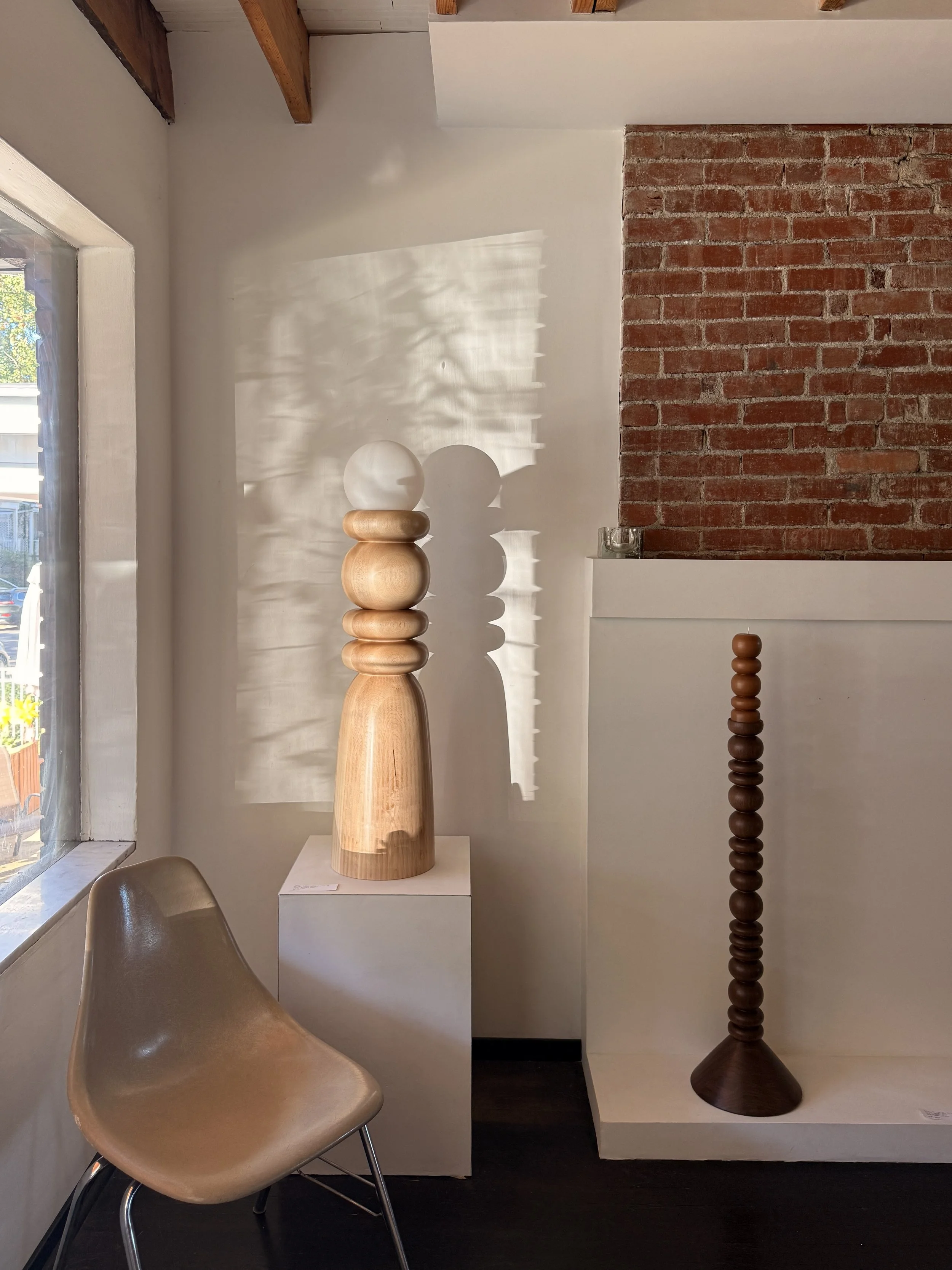 A modern interior with a beige chair near a window, a white pedestal with a wooden sculpture, and a tall wooden stand, against a wall with a brick section and a white section with shadows.
