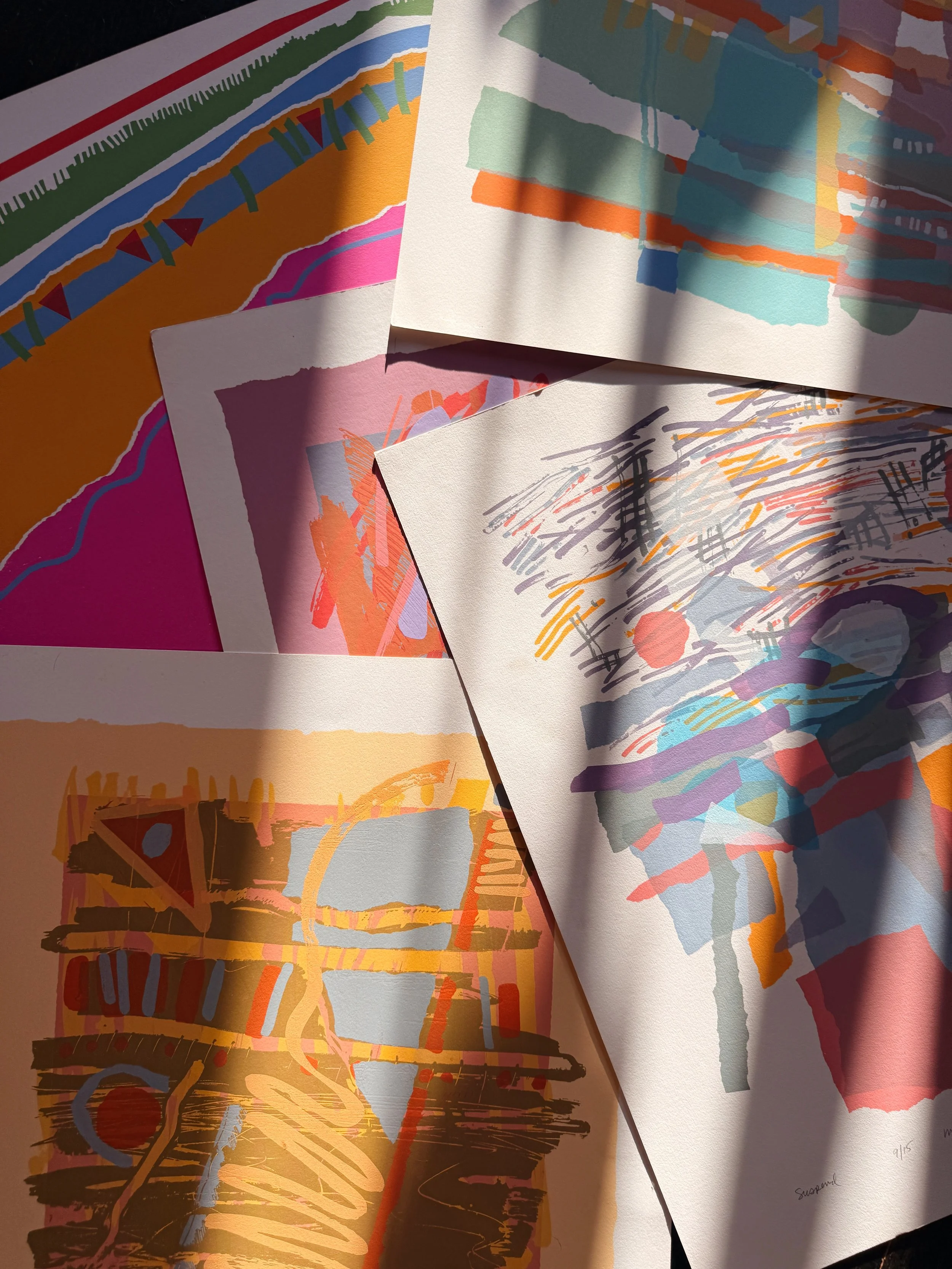 Several colorful abstract paintings and drawings with vibrant colors on sheets of paper. Shadows of window blinds cast over the artwork.