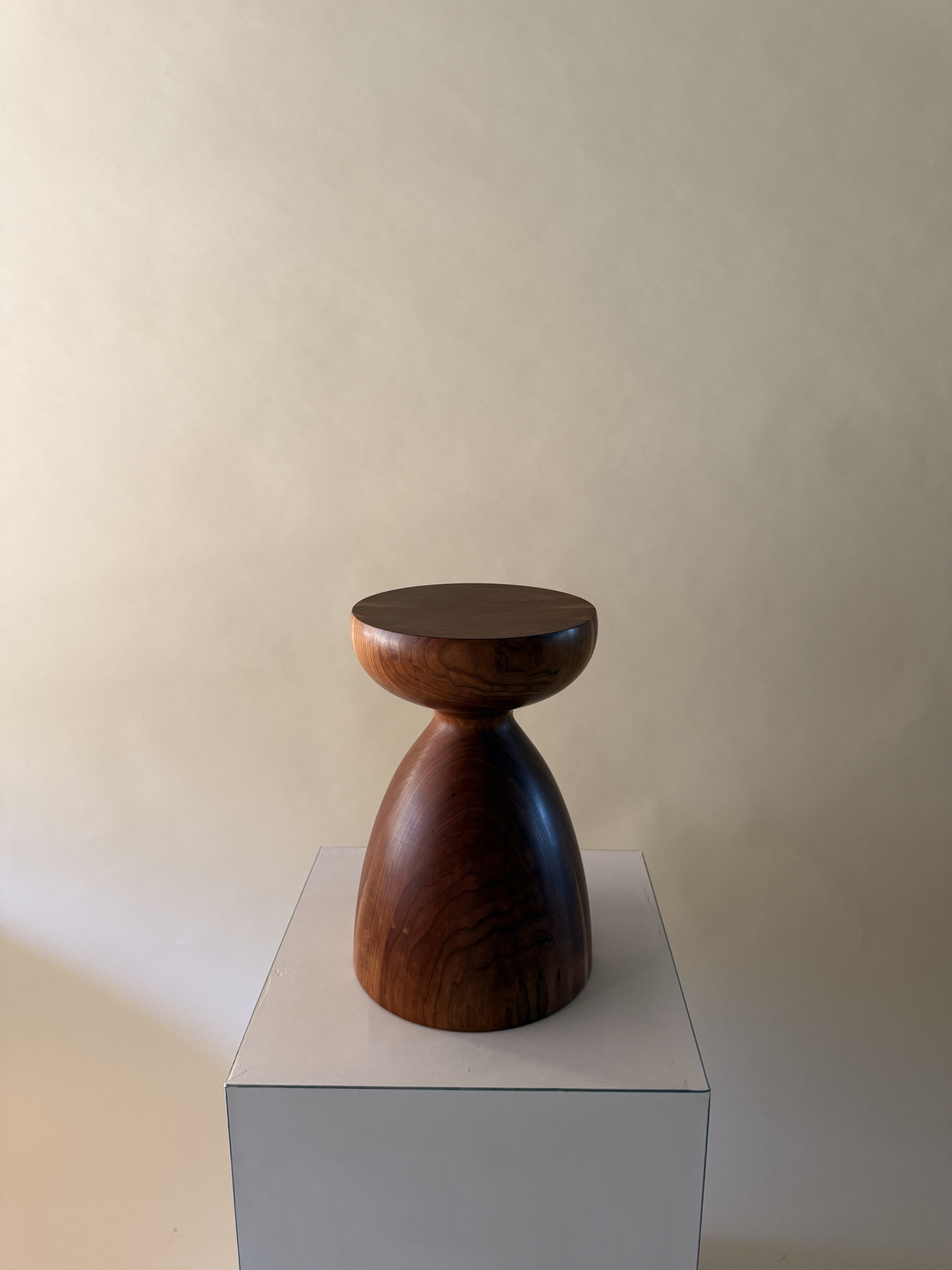 A wooden sculpture with a rounded top and a curved, elongated base, displayed on a white pedestal against a plain wall.