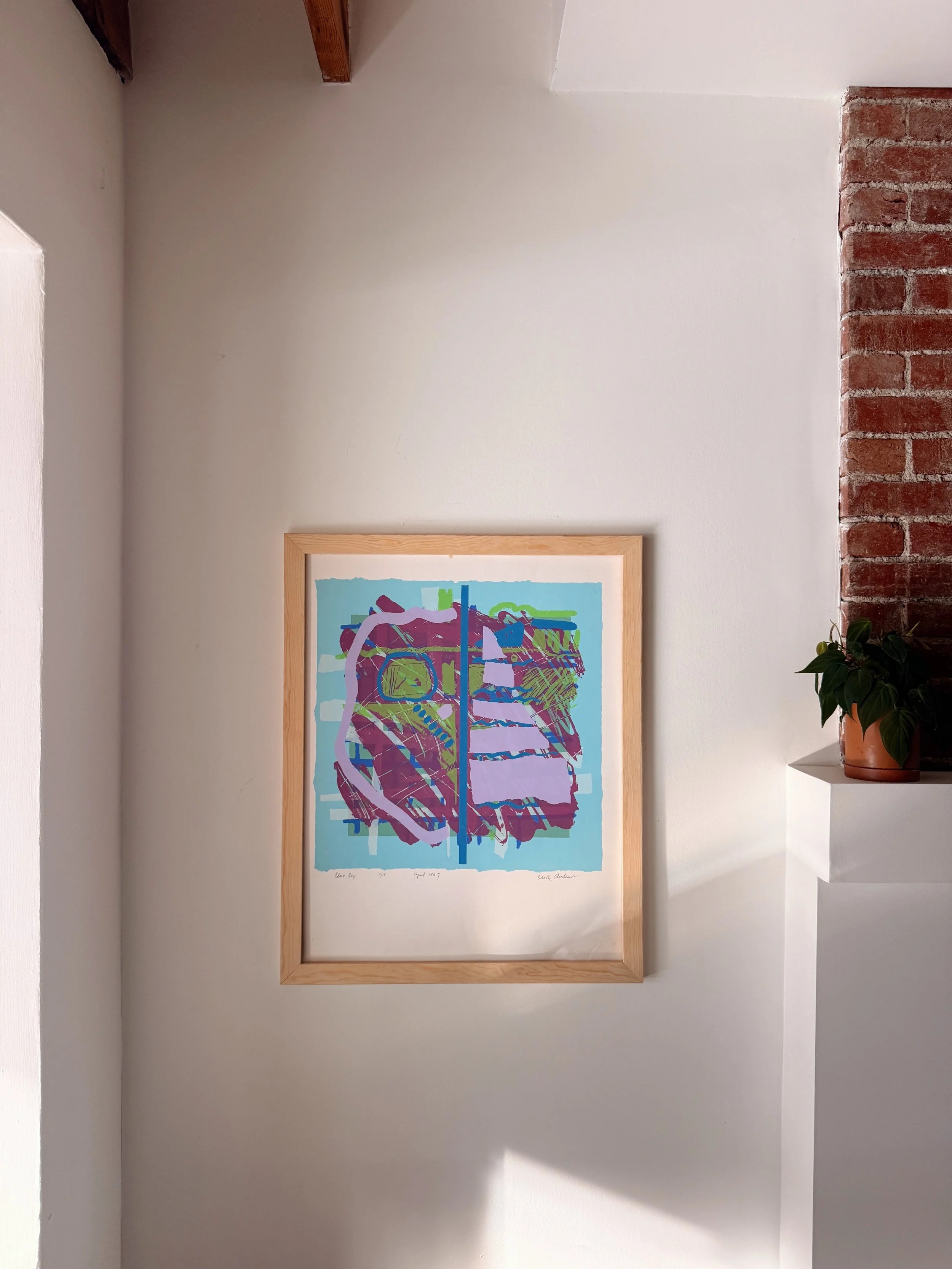 A framed abstract artwork hung on a white wall, with a small green plant in a terracotta pot on a white shelf to the right.