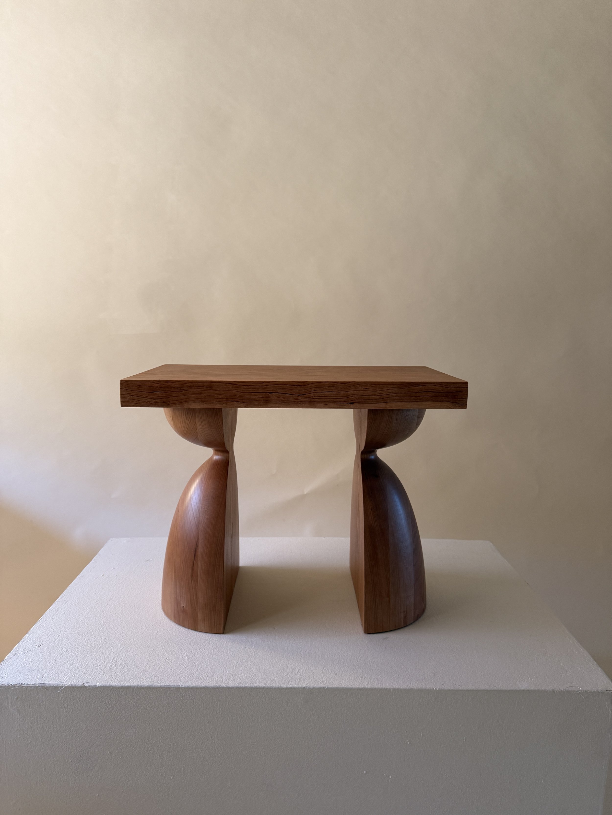 A small wooden table with a flat rectangular top and curved, sculptural legs, displayed on a white platform against a beige wall.