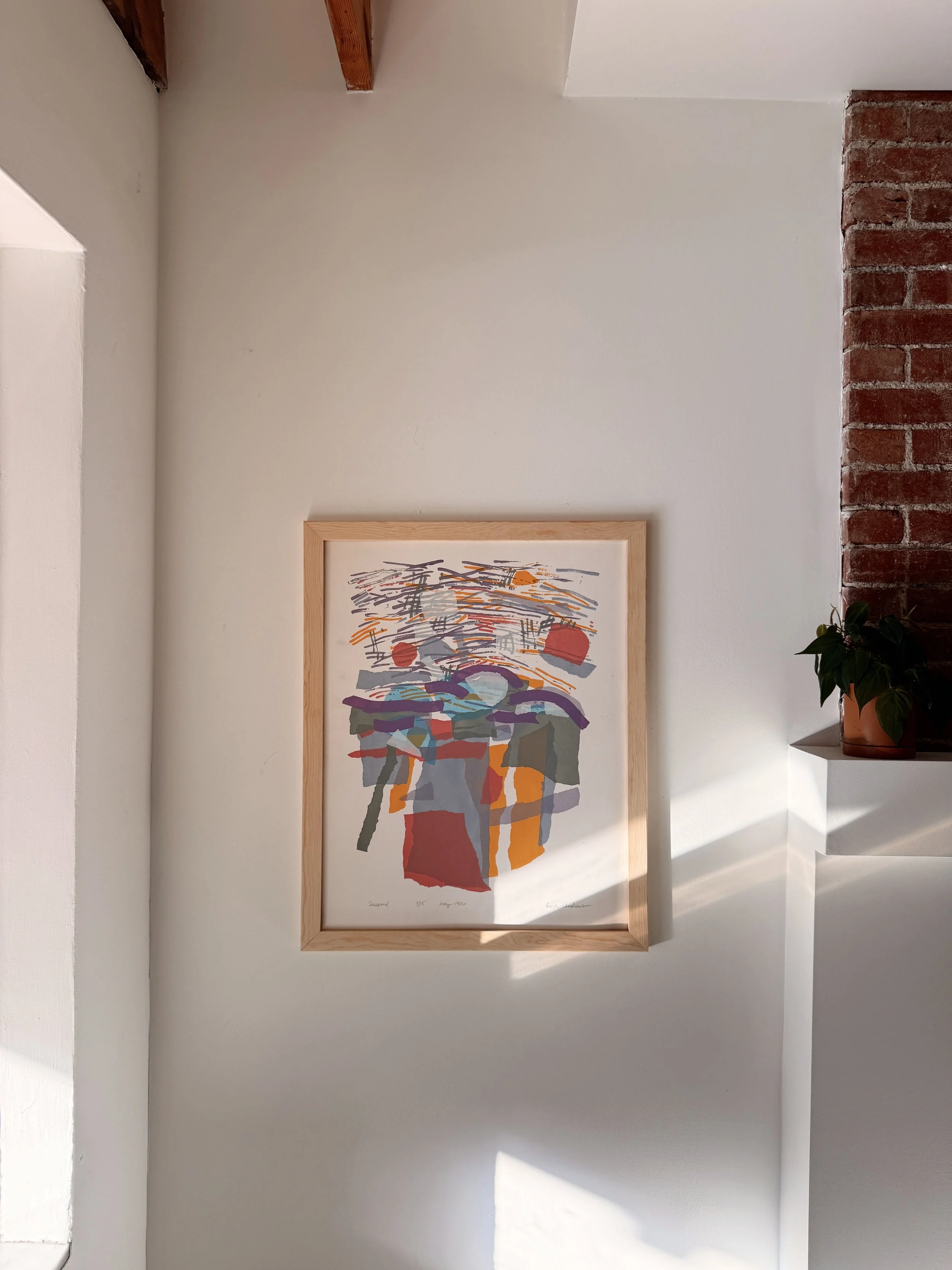 Framed abstract artwork hanging on a white wall in a room with a brick column and a potted plant in the corner.