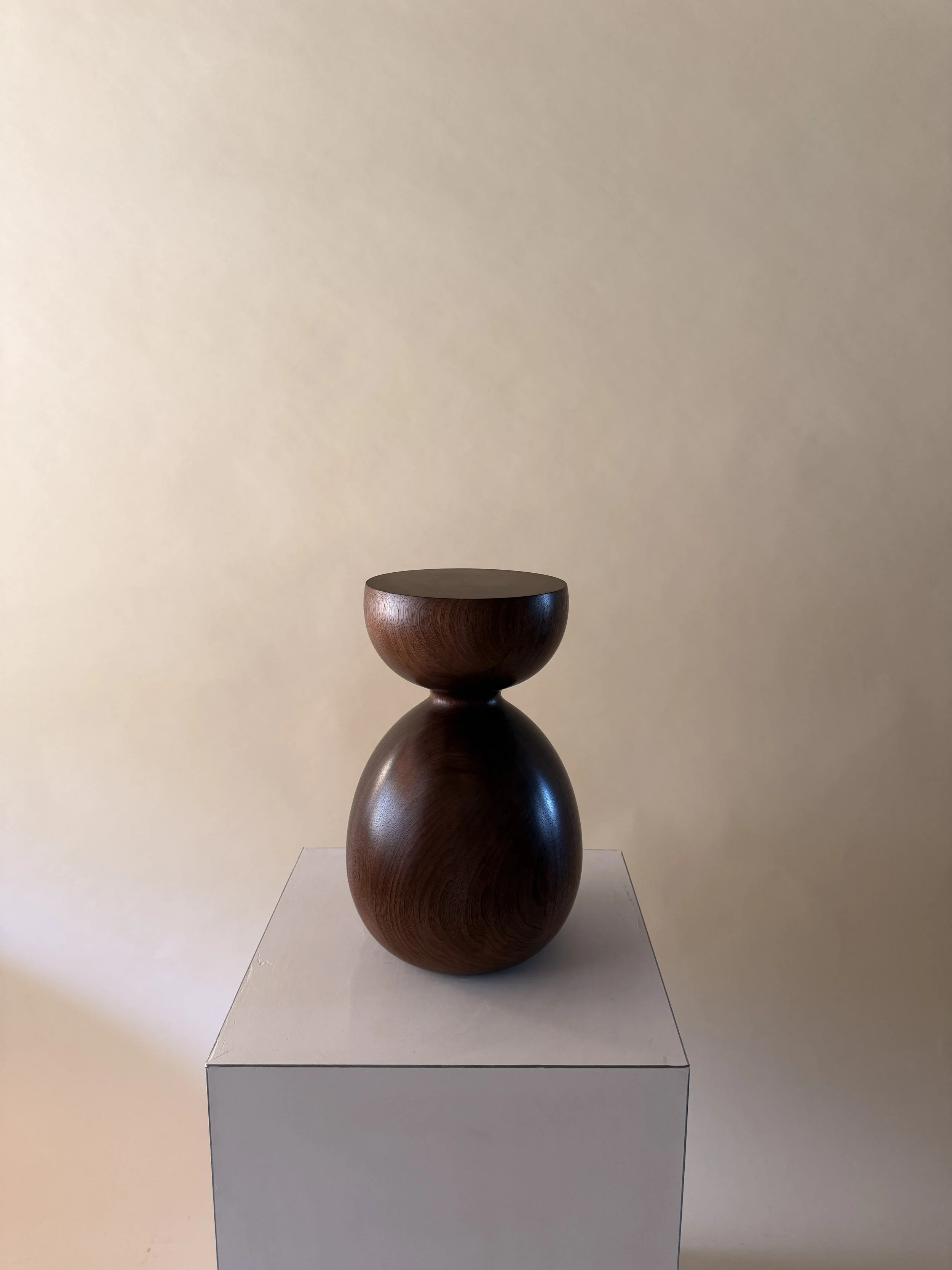 A wooden sculpture consisting of three stacked, rounded, and hollow shapes on a white pedestal against a neutral wall.
