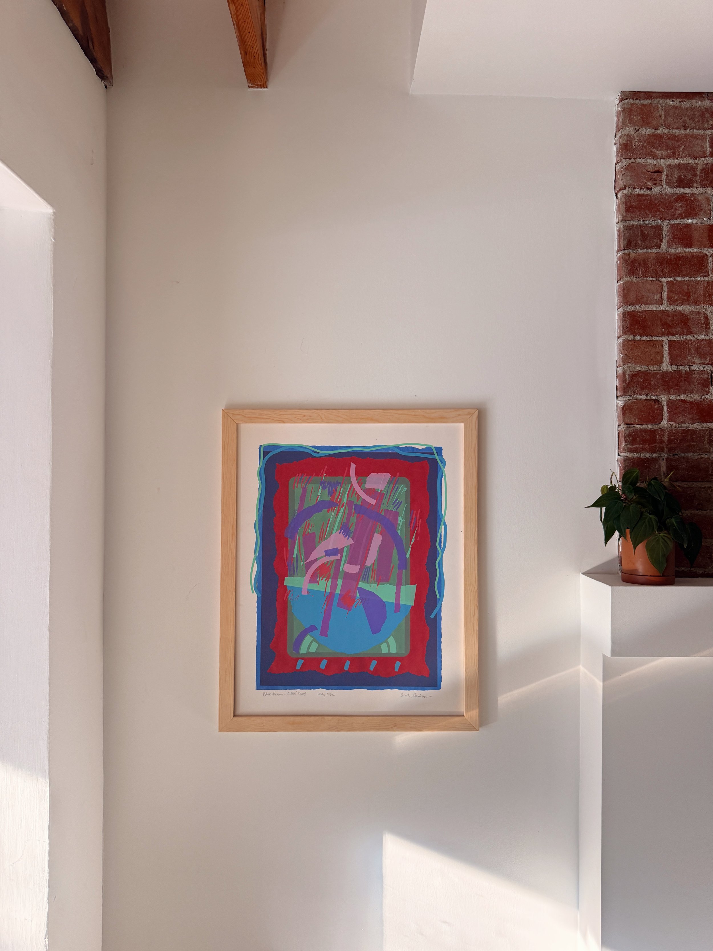 Colorful abstract artwork framed on a white wall, next to a brick column and a potted plant.