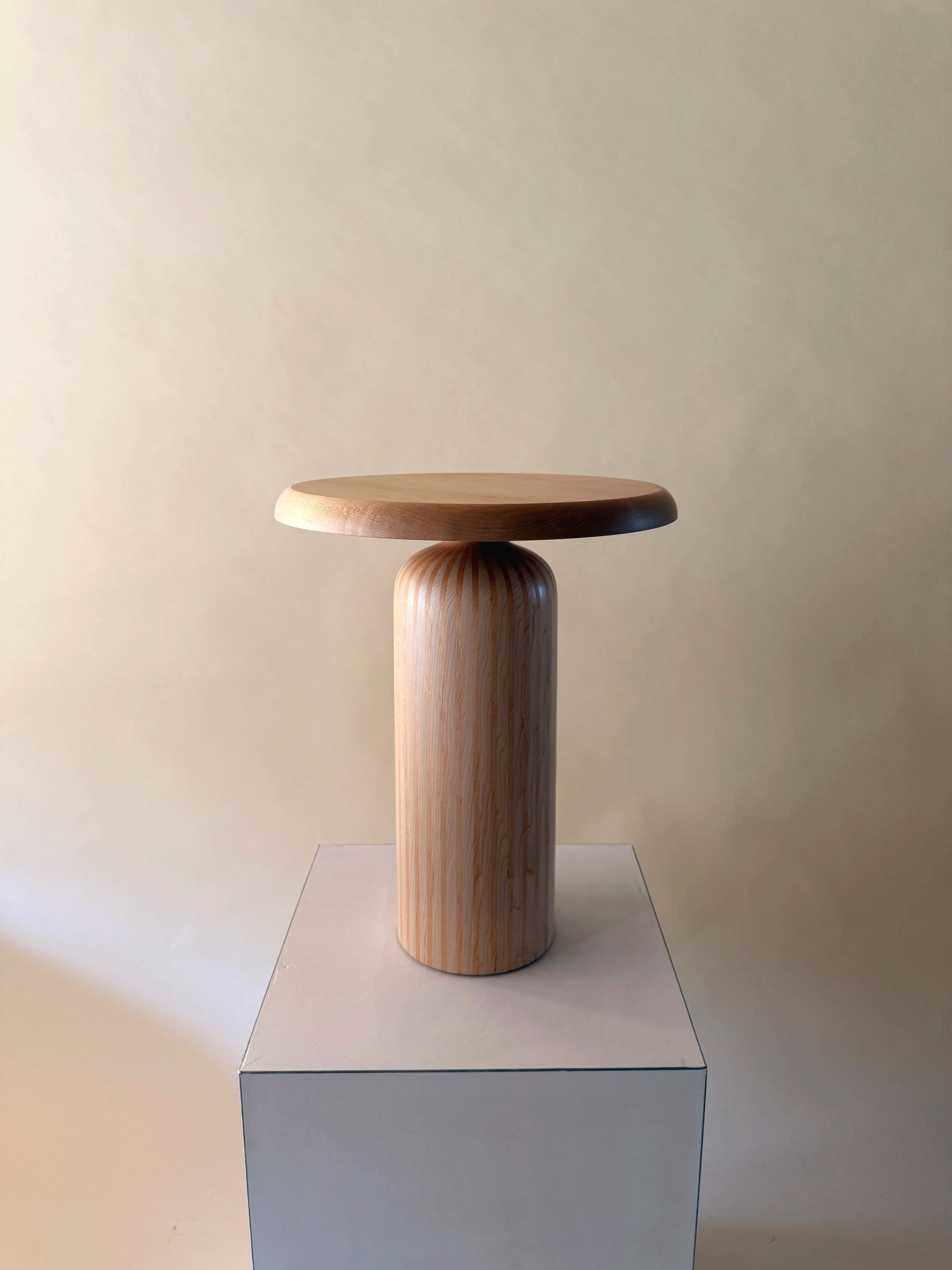 A wooden stool with a round seat and cylindrical base, placed on a white platform against a plain beige wall.