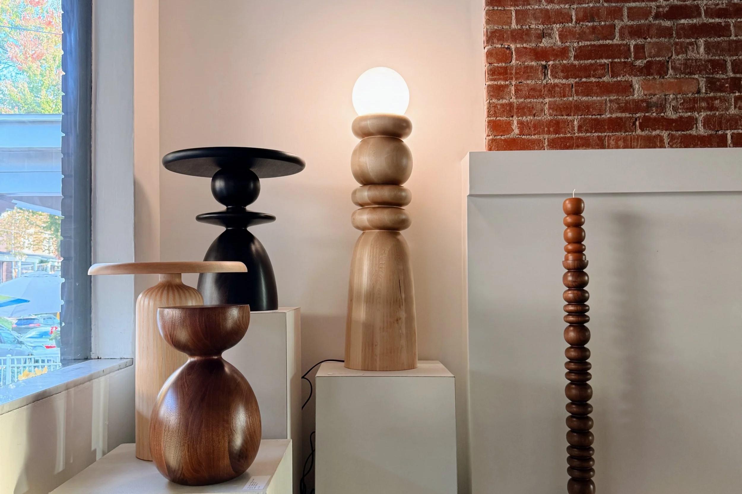Various wooden sculptures on display in a gallery, with a window to the left and a brick wall to the right.