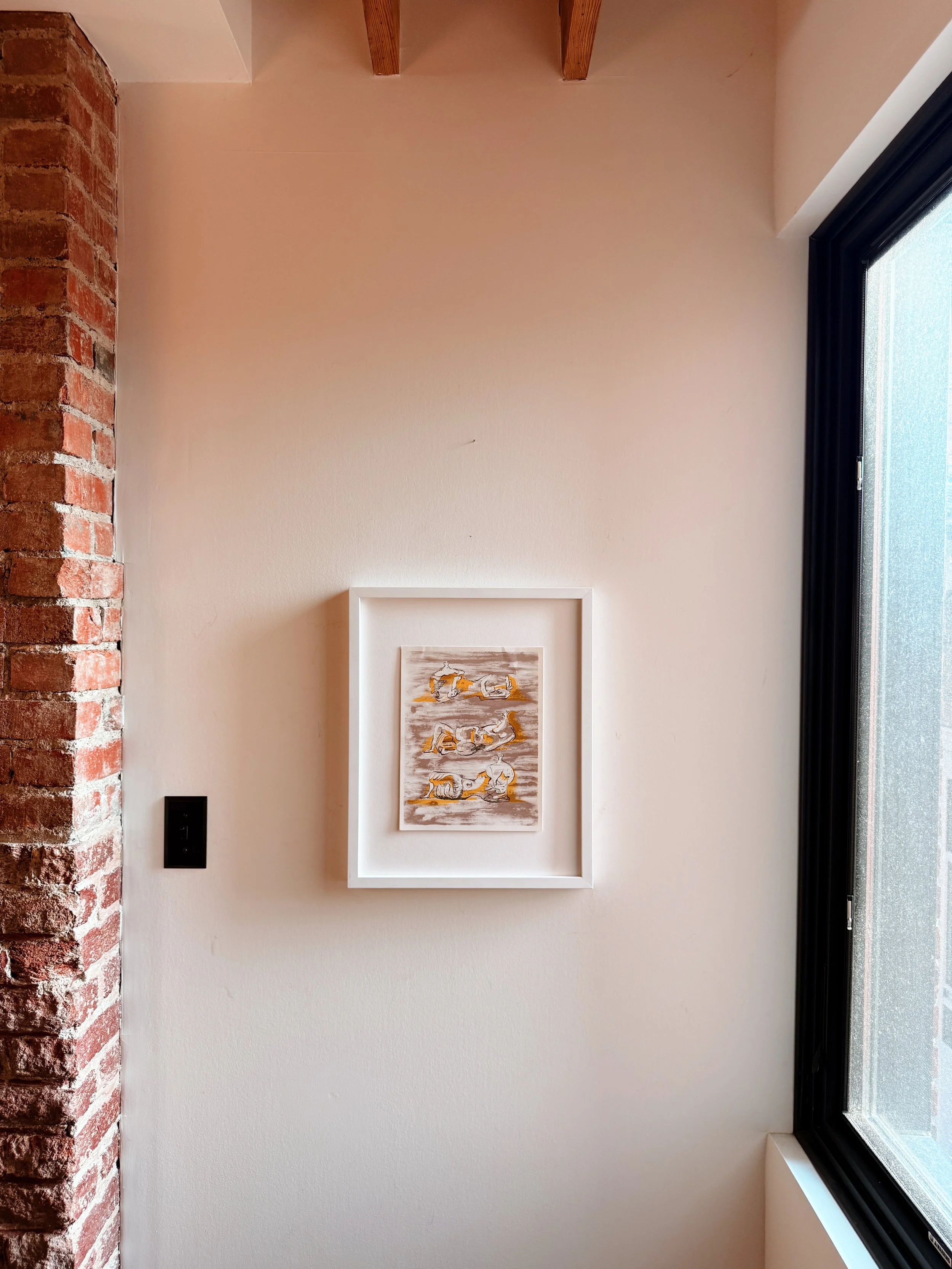 A framed artwork hanging on a beige wall next to a brick column and a large window with black framing. The artwork depicts abstract shapes resembling animals in shades of gray, white, and yellow.