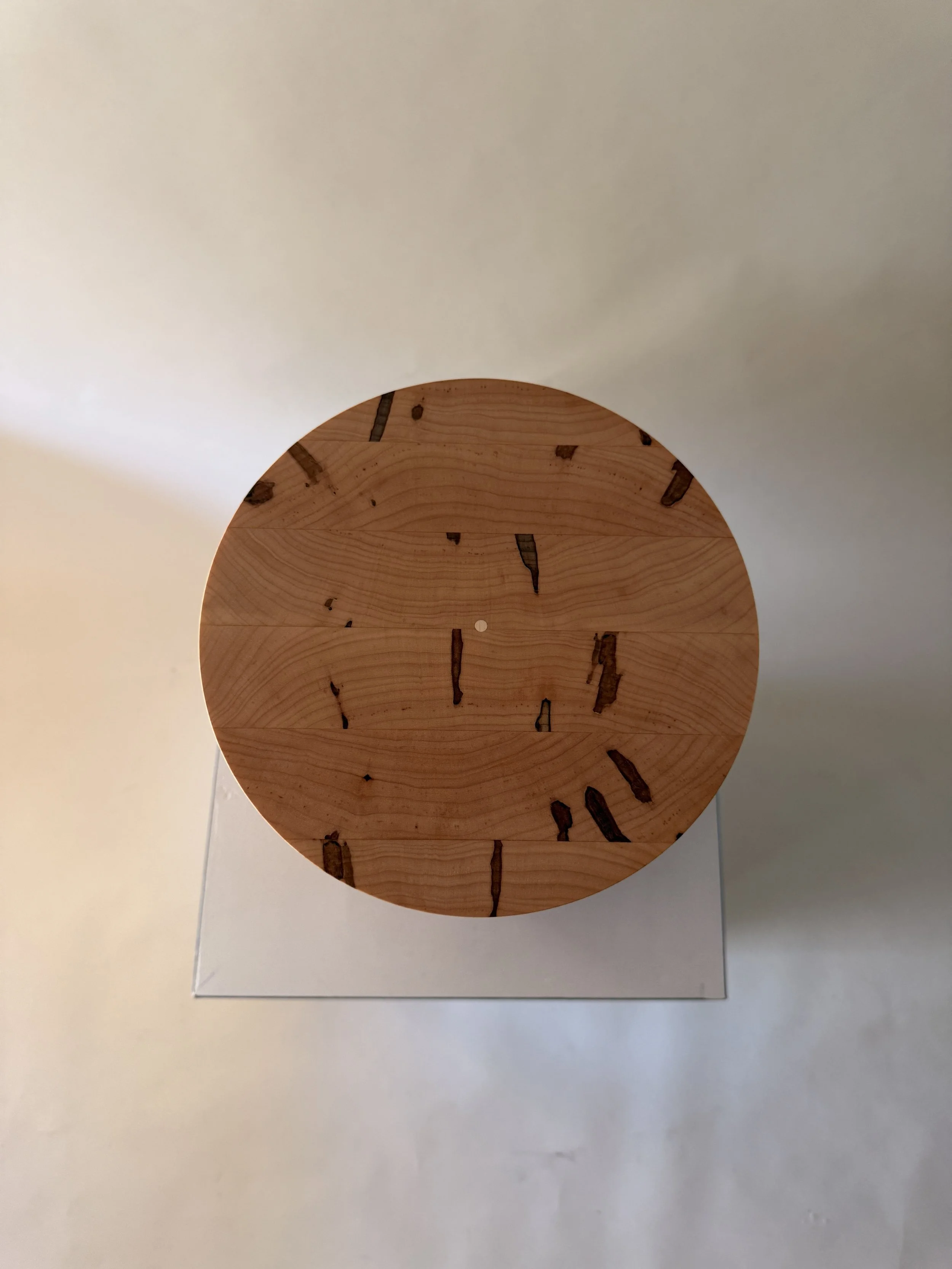 A round wooden clock with black Roman numerals and hands, placed on a white surface with a mirror behind it.