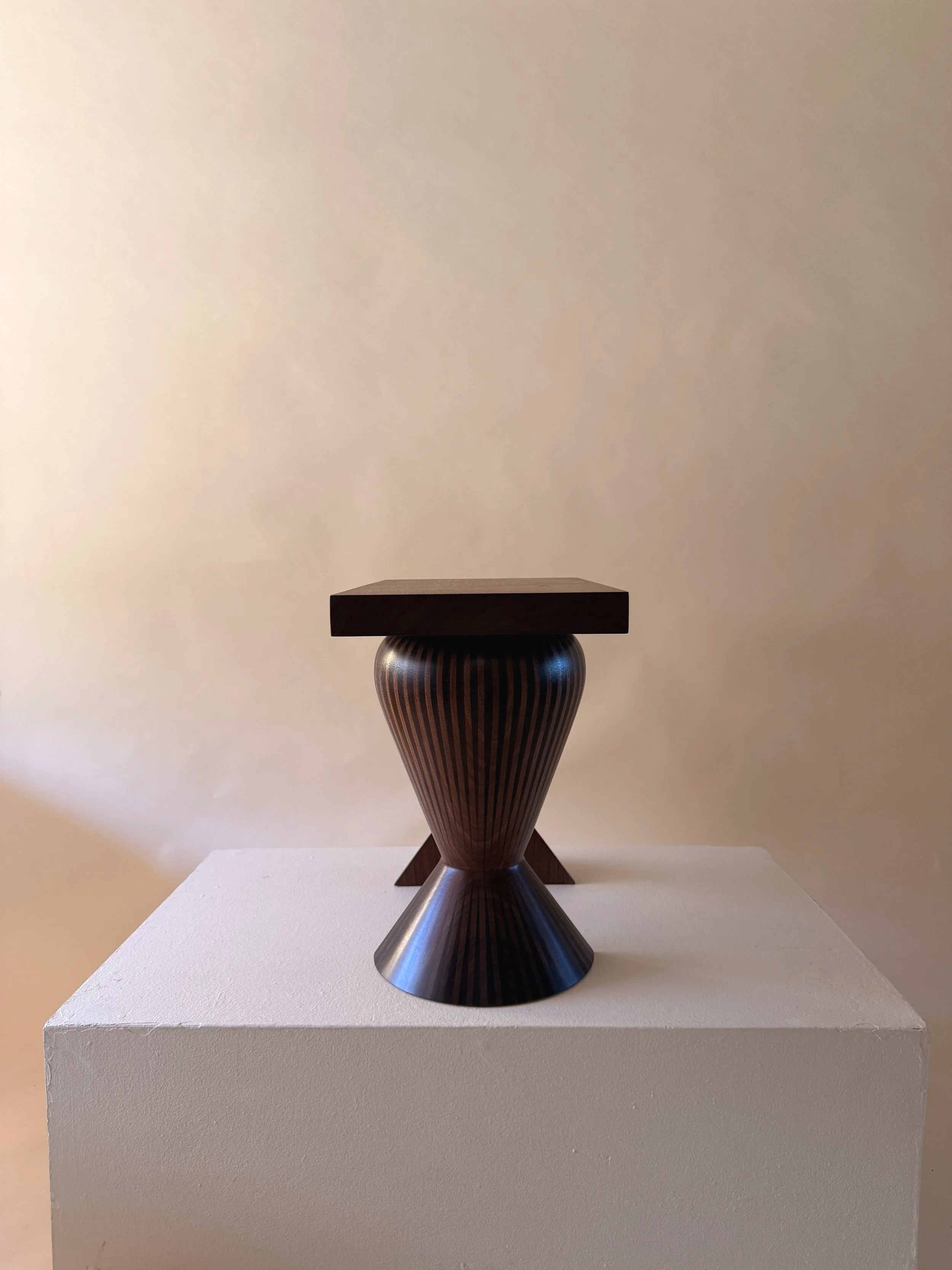 A decorative table with a wooden top and a patterned, cone-shaped metal base, displayed on a white pedestal against a plain beige wall.