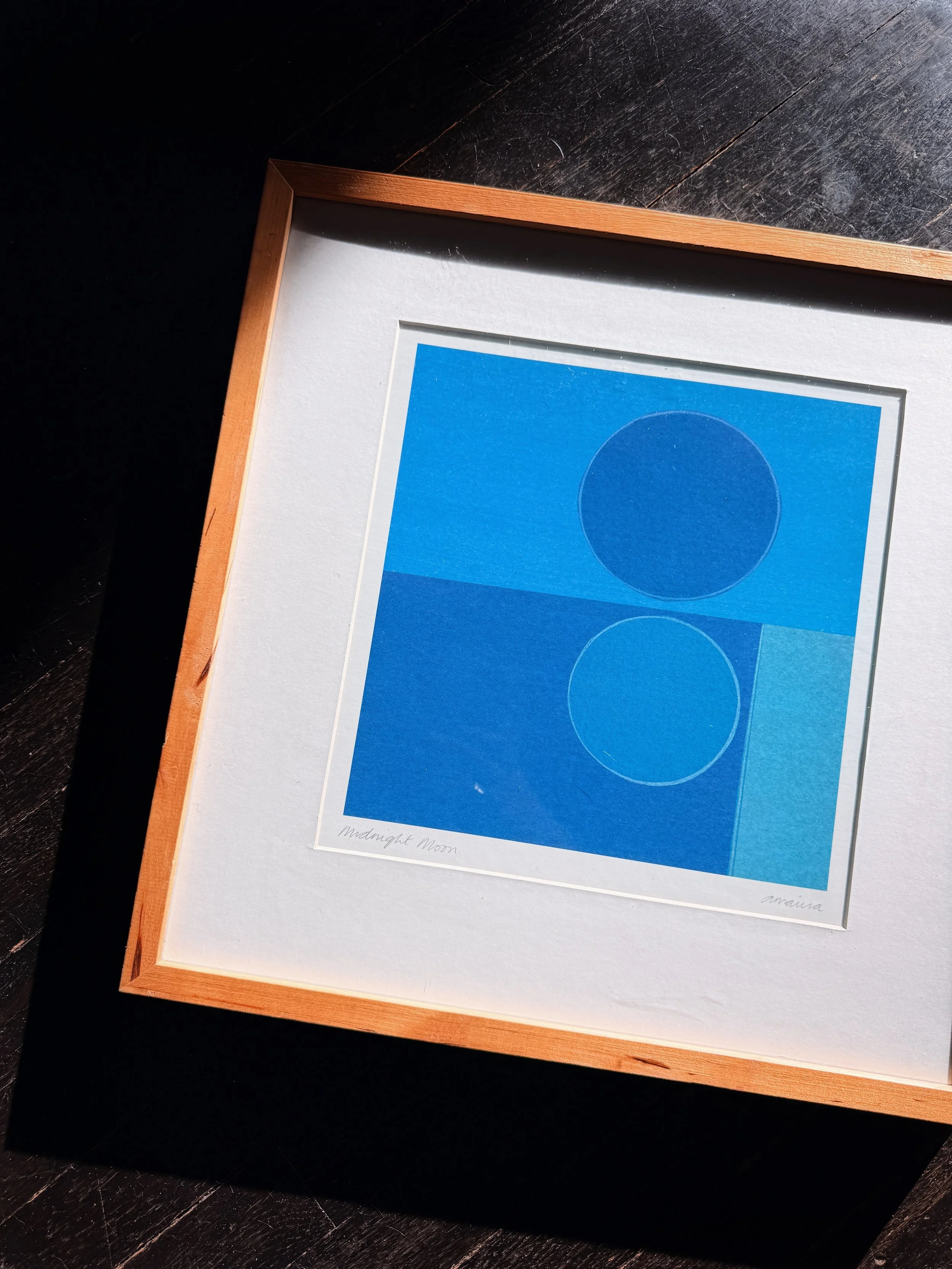 Framed blue artwork featuring circles on white matte, placed on black wooden surface with sunlight casting shadows.