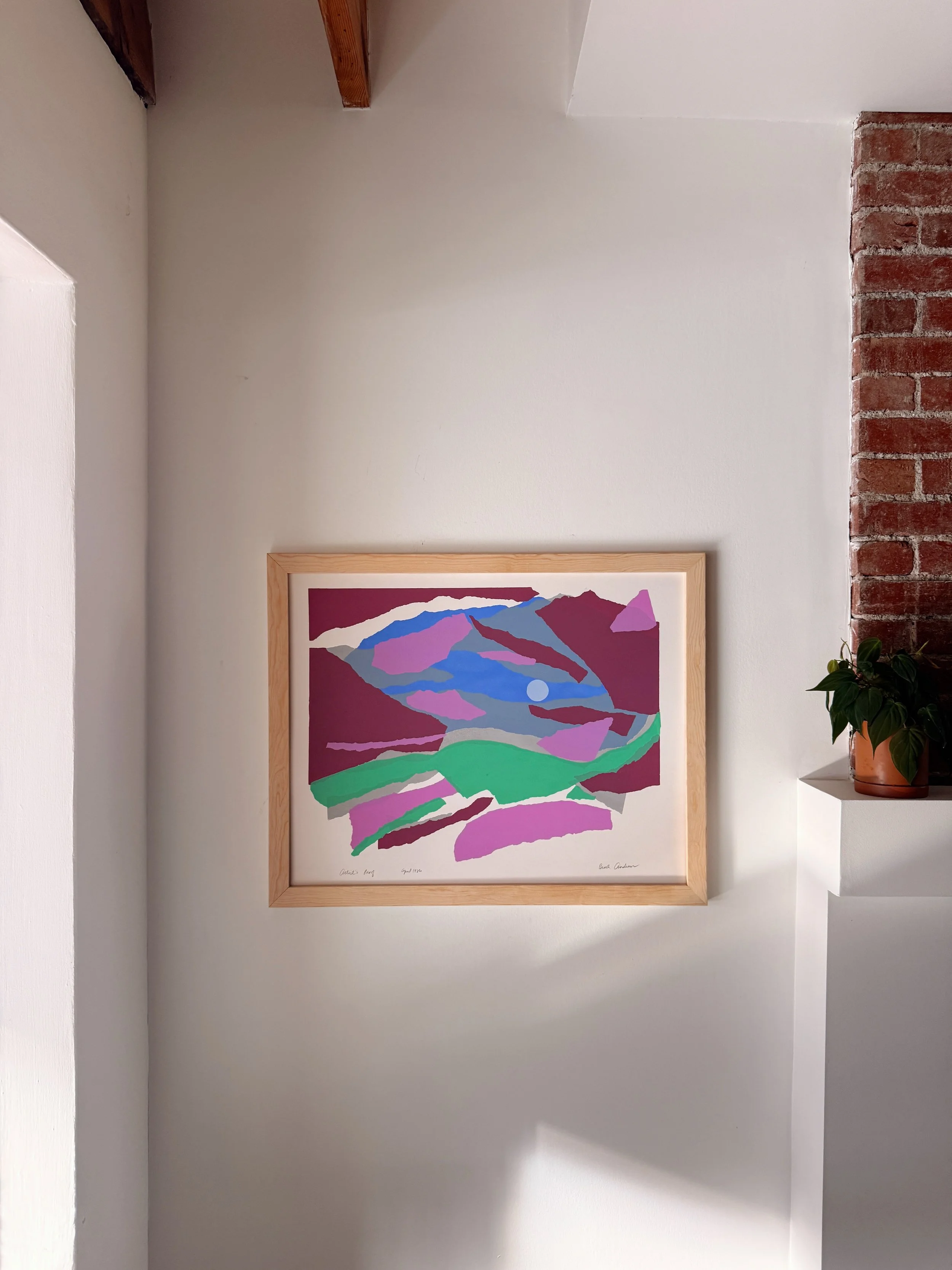 An art print of an abstract landscape painting with mountains, hills, and sky in shades of purple, pink, blue, green, and white, framed in light wood, hanging on a white wall near a brick column and a small plant with green leaves in a terracotta pot.