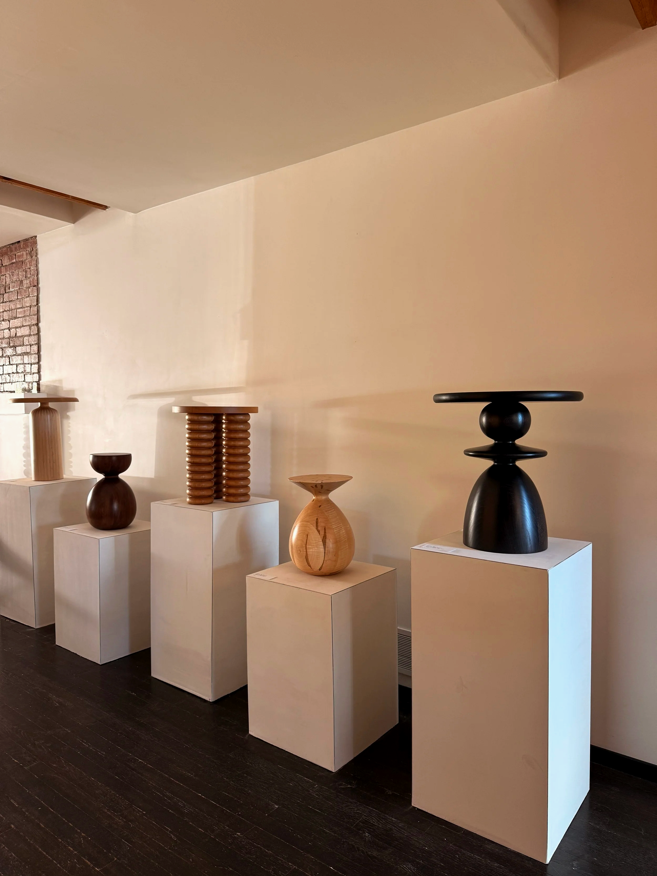 Various wooden sculptures displayed on white pedestals in an art gallery, with a brick wall in the background and a dark wooden floor.