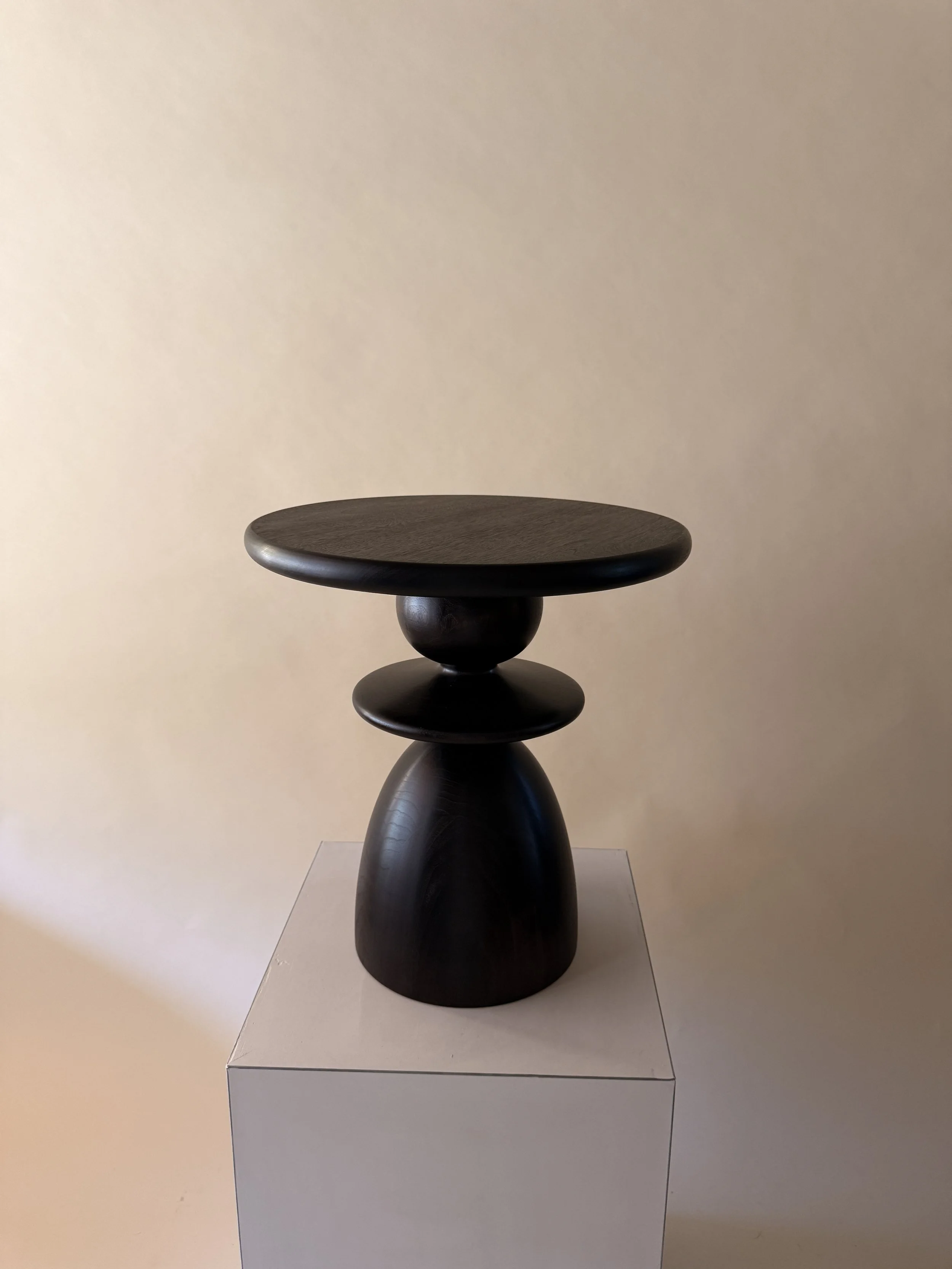A dark wooden pedestal table with a rounded top and a sculptural, rounded base, placed on a white pedestal against a beige wall.