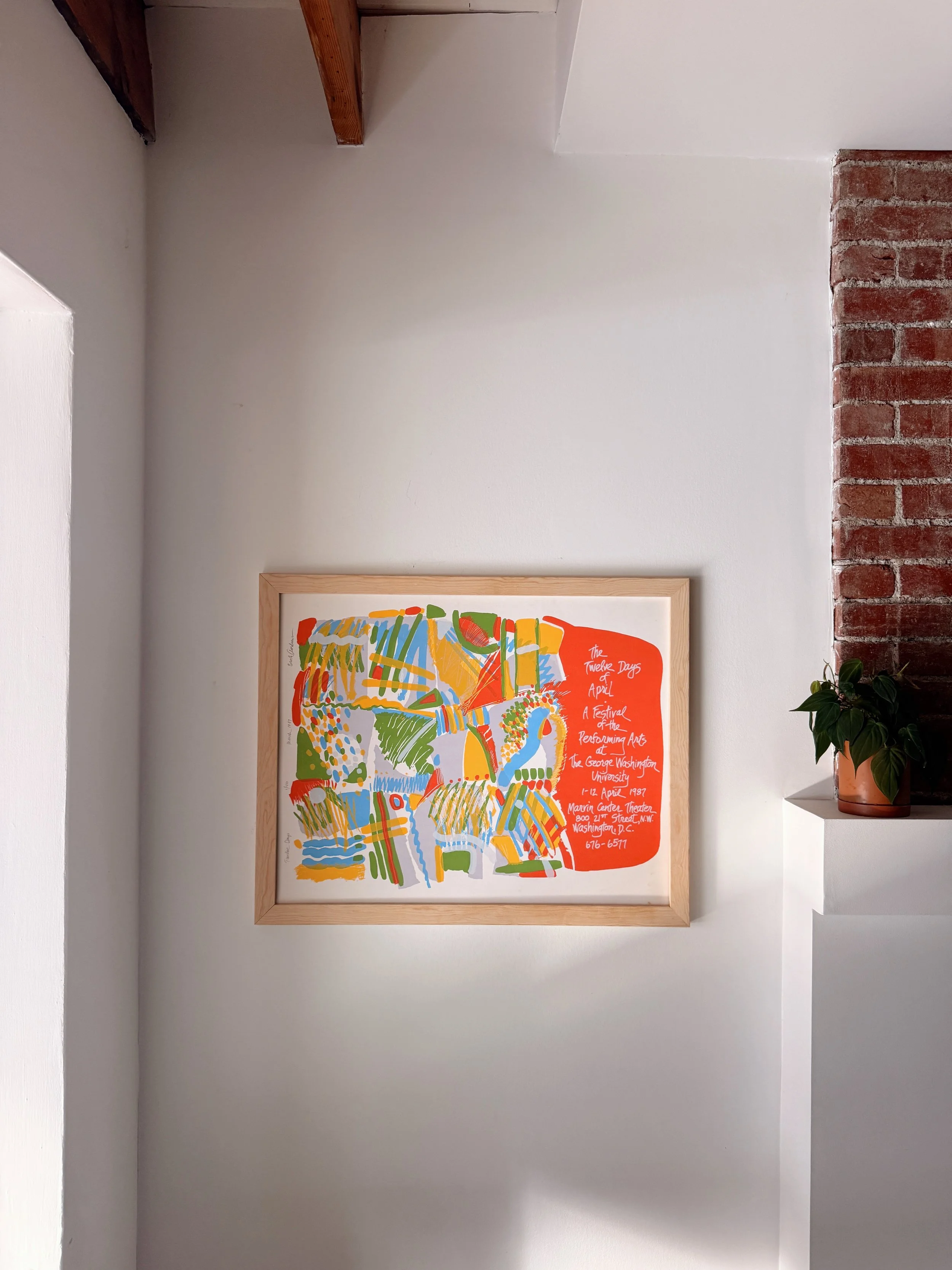 A colorful poster with abstract artwork and text about an event at George Washington University in April 1987, hanging on a white wall with a brick section on the right, a potted plant on a white shelf, and a wooden beam on the ceiling.