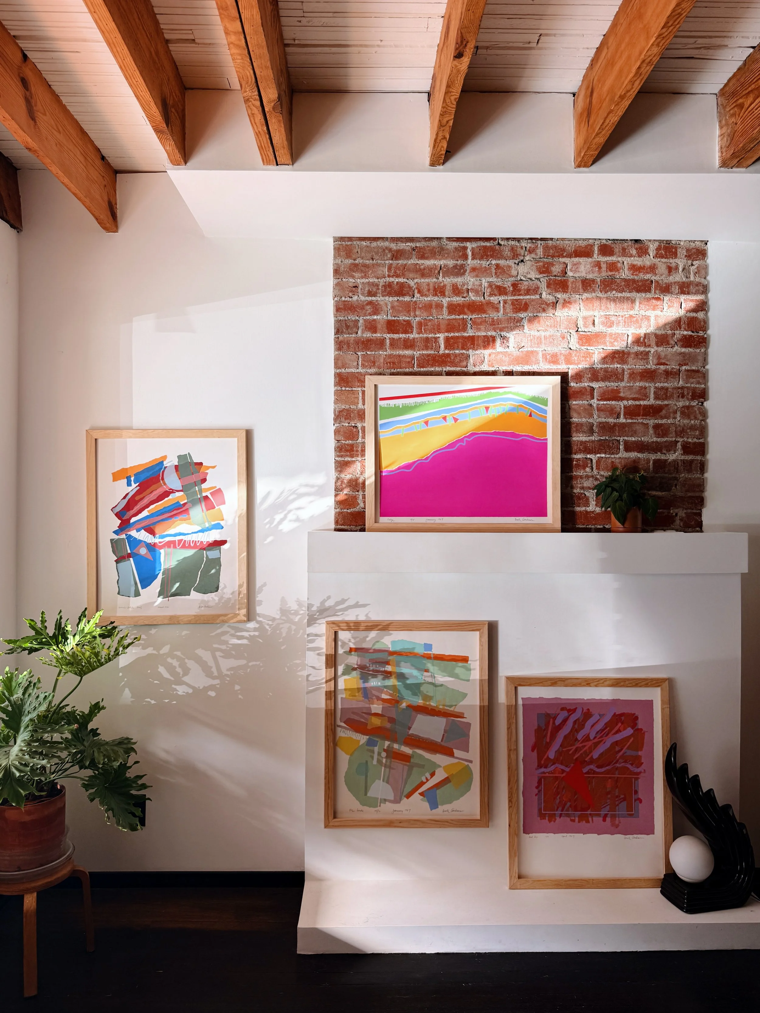 Gallery wall with colorful abstract artwork, exposed brick accent wall, and wooden beams ceiling. Includes a potted plant and a black object on a white shelf.