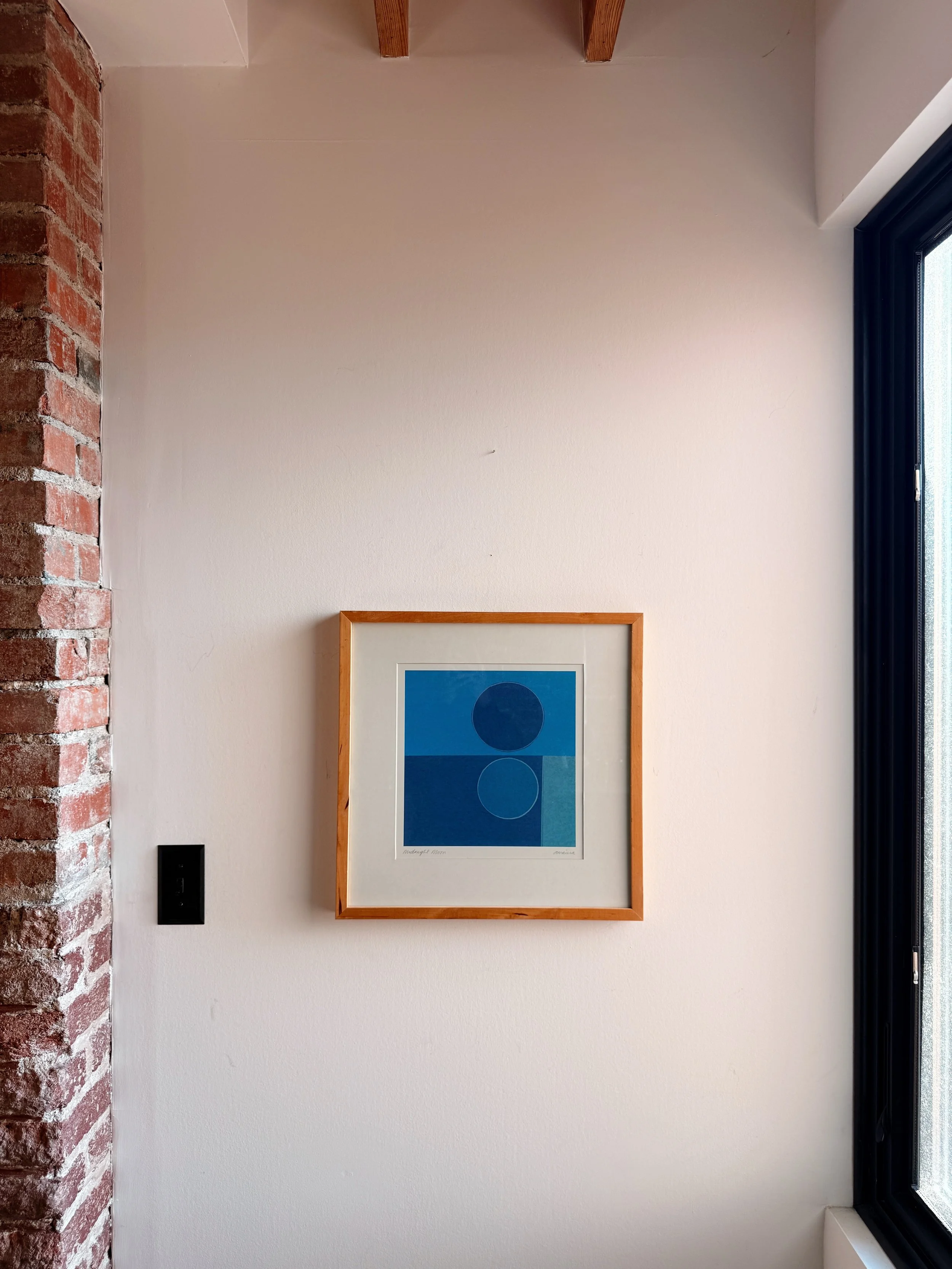 Interior wall with framed abstract artwork featuring blue circles, a brick wall on the left, a window with black trim on the right, and ceiling beams.