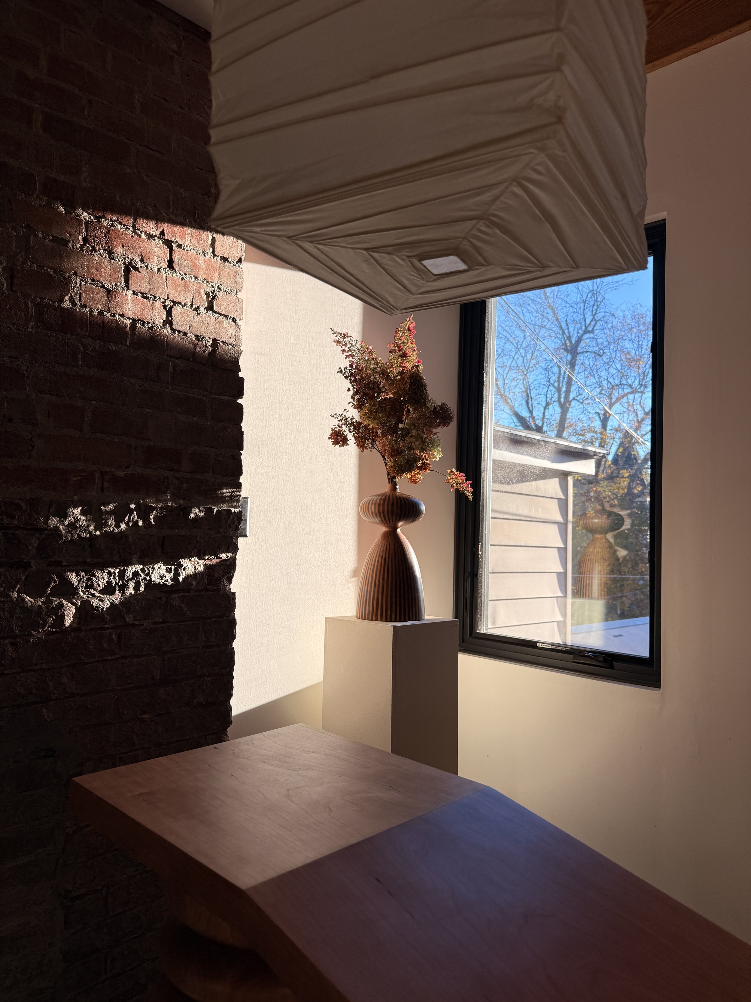 Interior scene with a wooden table, a brick wall, a window showing trees and a building outside, a vase with dried flowers, and a suspended fabric covering, likely a lampshade or canopy.