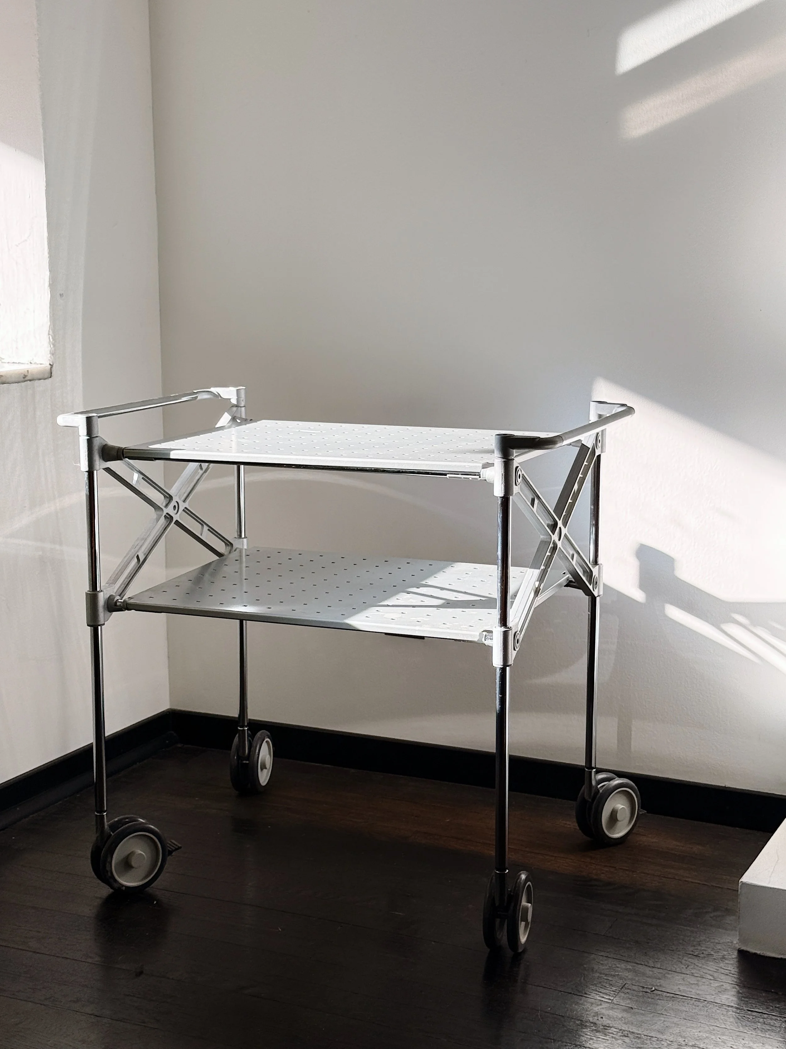 A white medical or hospital cart with two shelves, four wheels, and side rails, standing against a white wall nearby a window with sunlight casting shadows.