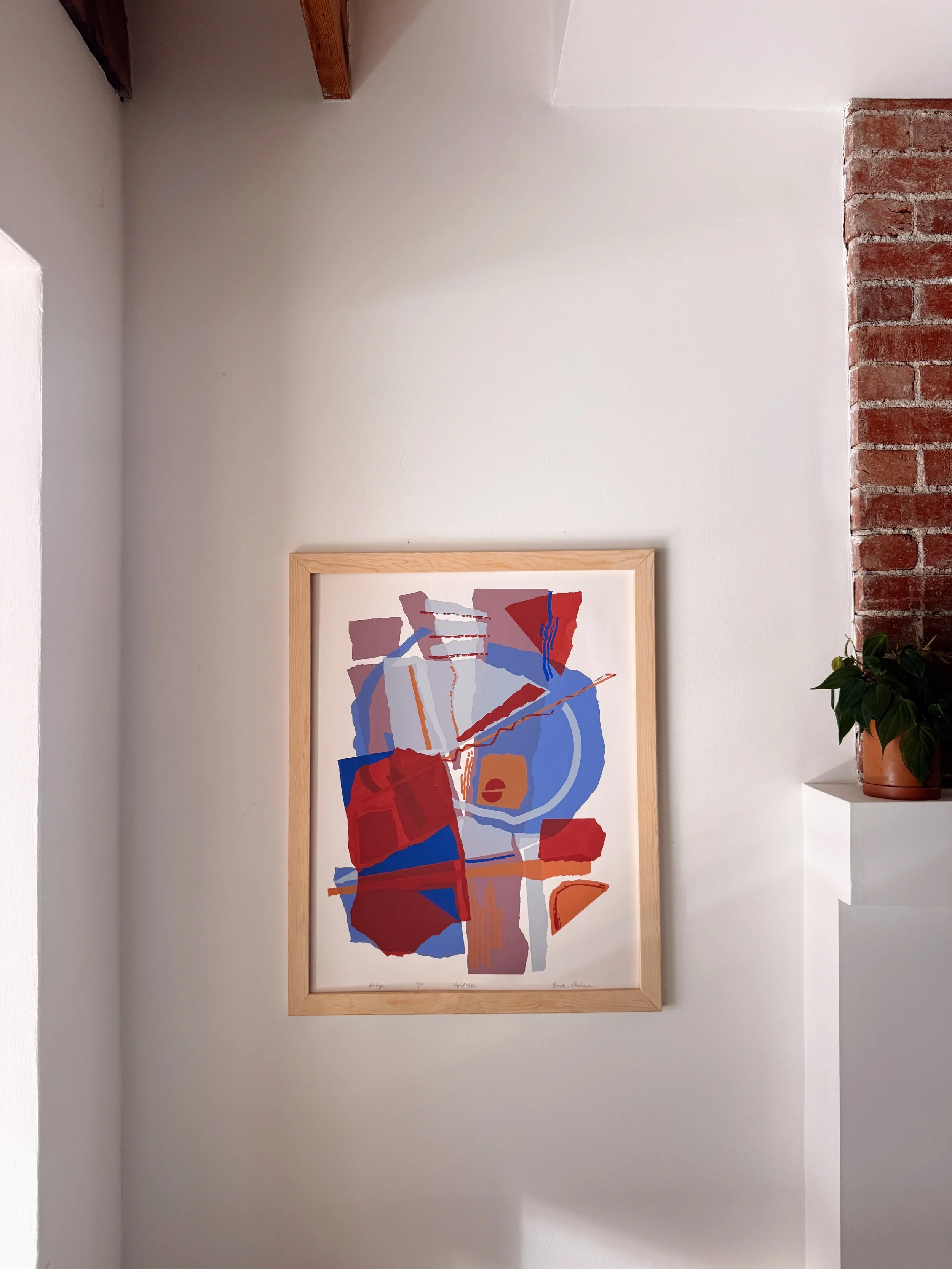 Colorful abstract art painting with geometric shapes in red, blue, pink, and orange, framed and hanging on a white wall next to a brick wall with a potted plant on a white shelf.