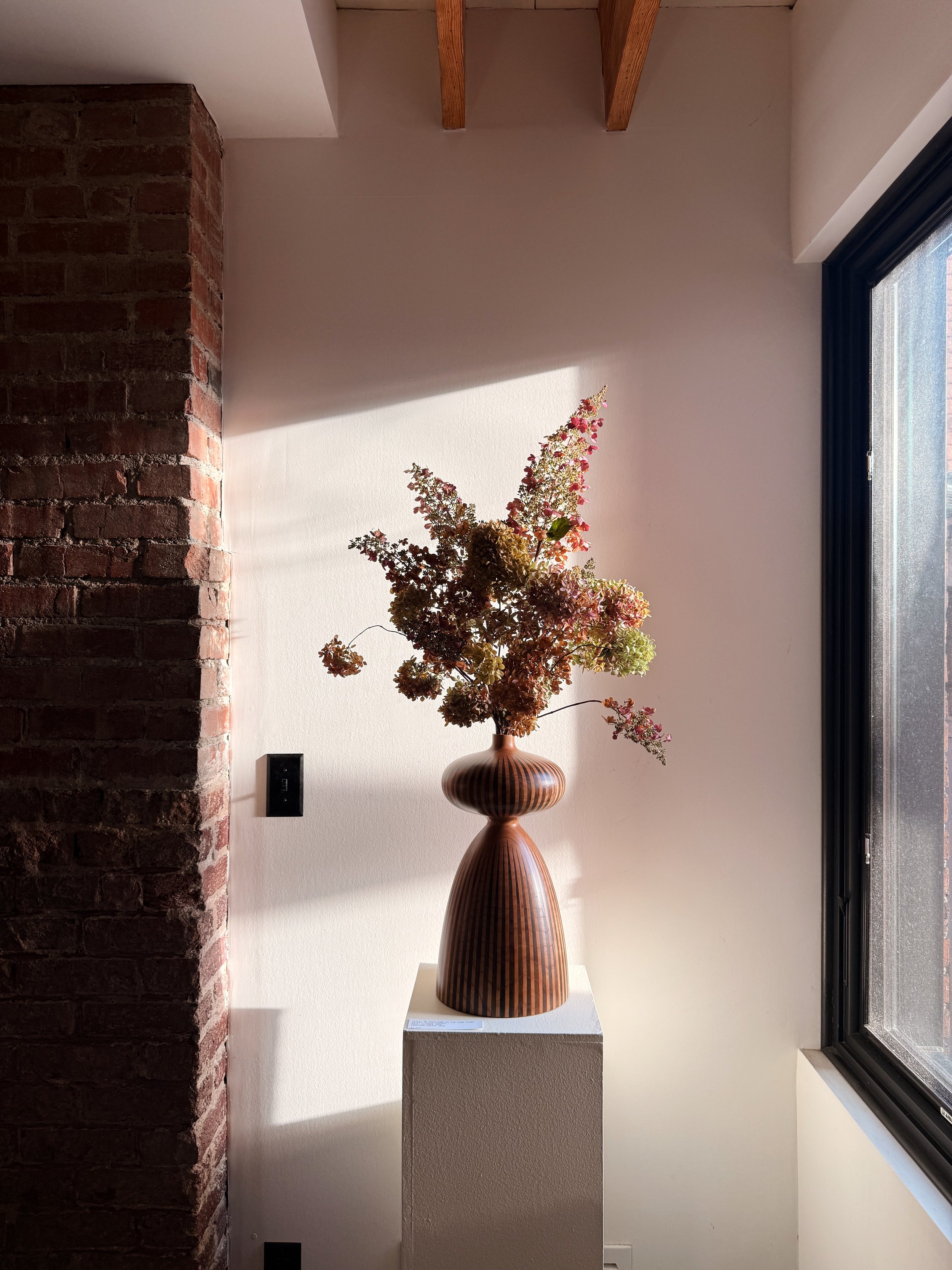 A decorative wooden vase with dried flowers inside, placed on a pedestal near a window with black frames and a white wall, illuminated by natural sunlight.