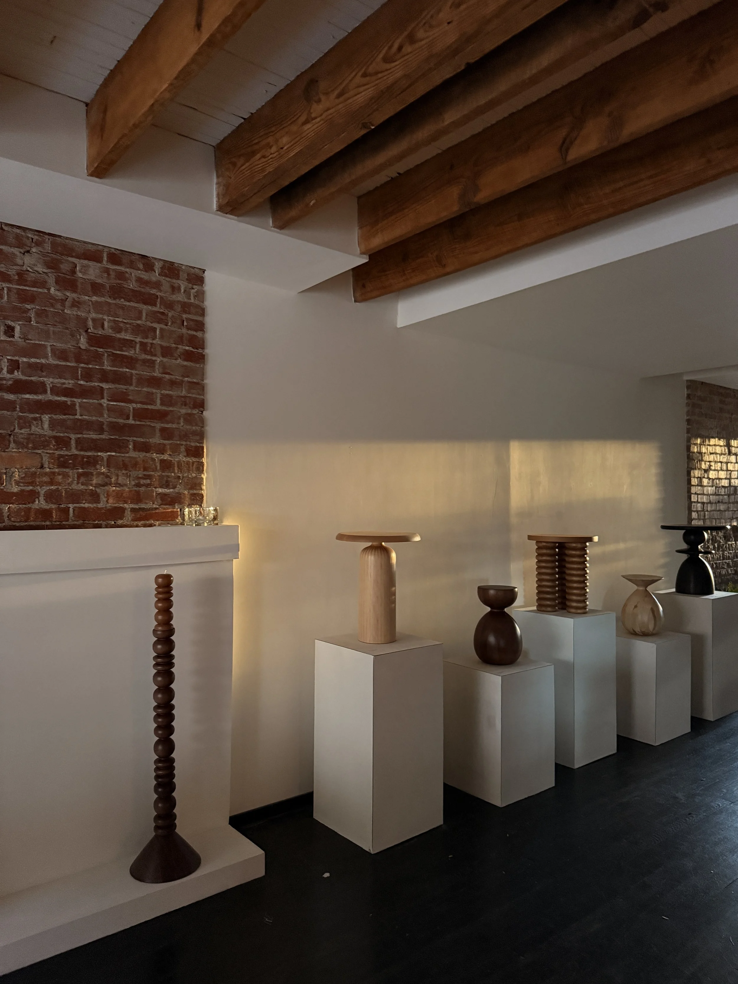 Display of wooden vases and sculptures on white pedestals inside a gallery space with brick and plaster walls.