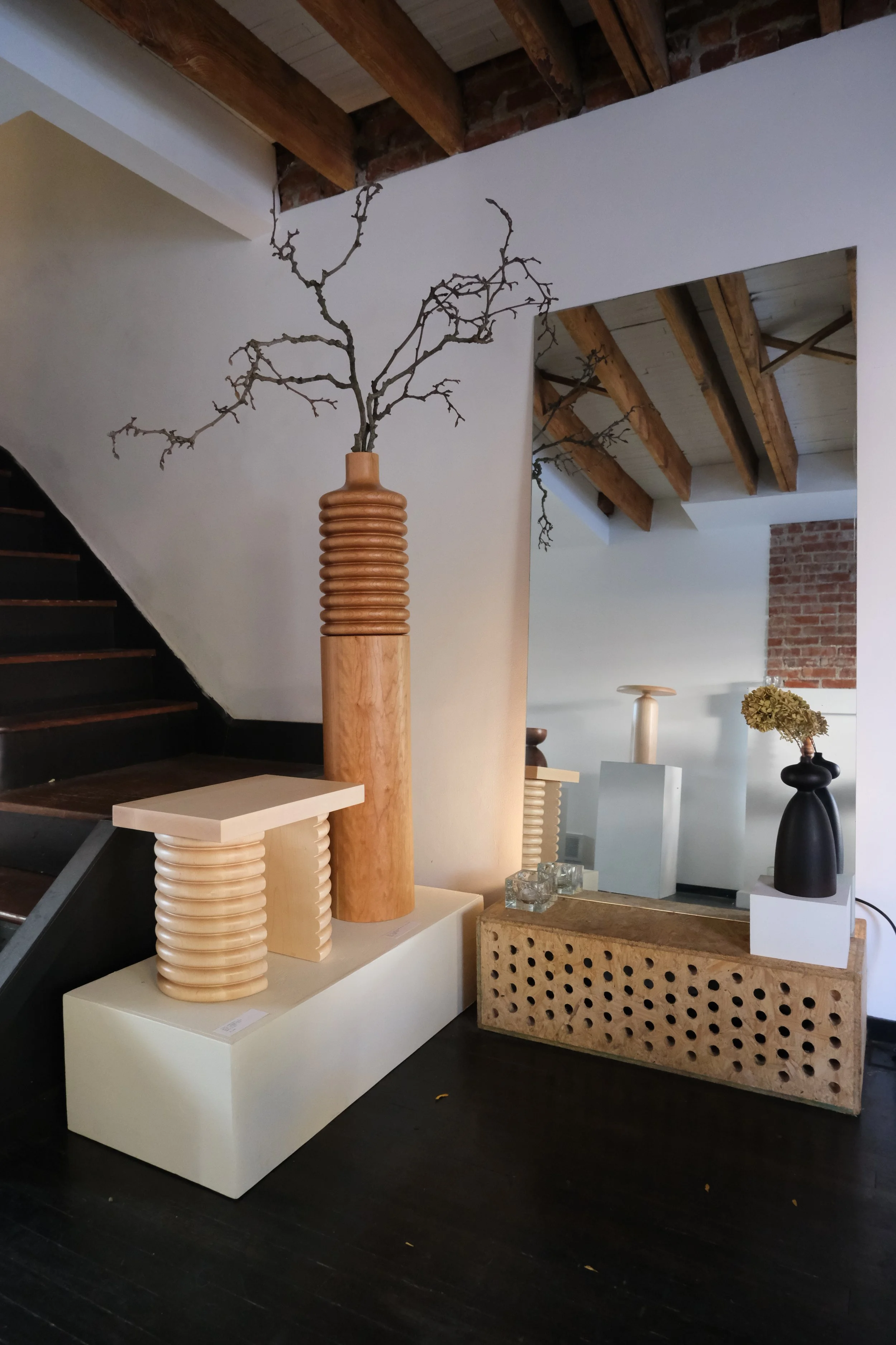 Interior view of a furniture and decor display, featuring a tall wooden vase with a leafless branch, a ribbed wooden sculpture, and various vases and tables in a room with dark hardwood floors, exposed roof beams, and brick accents.