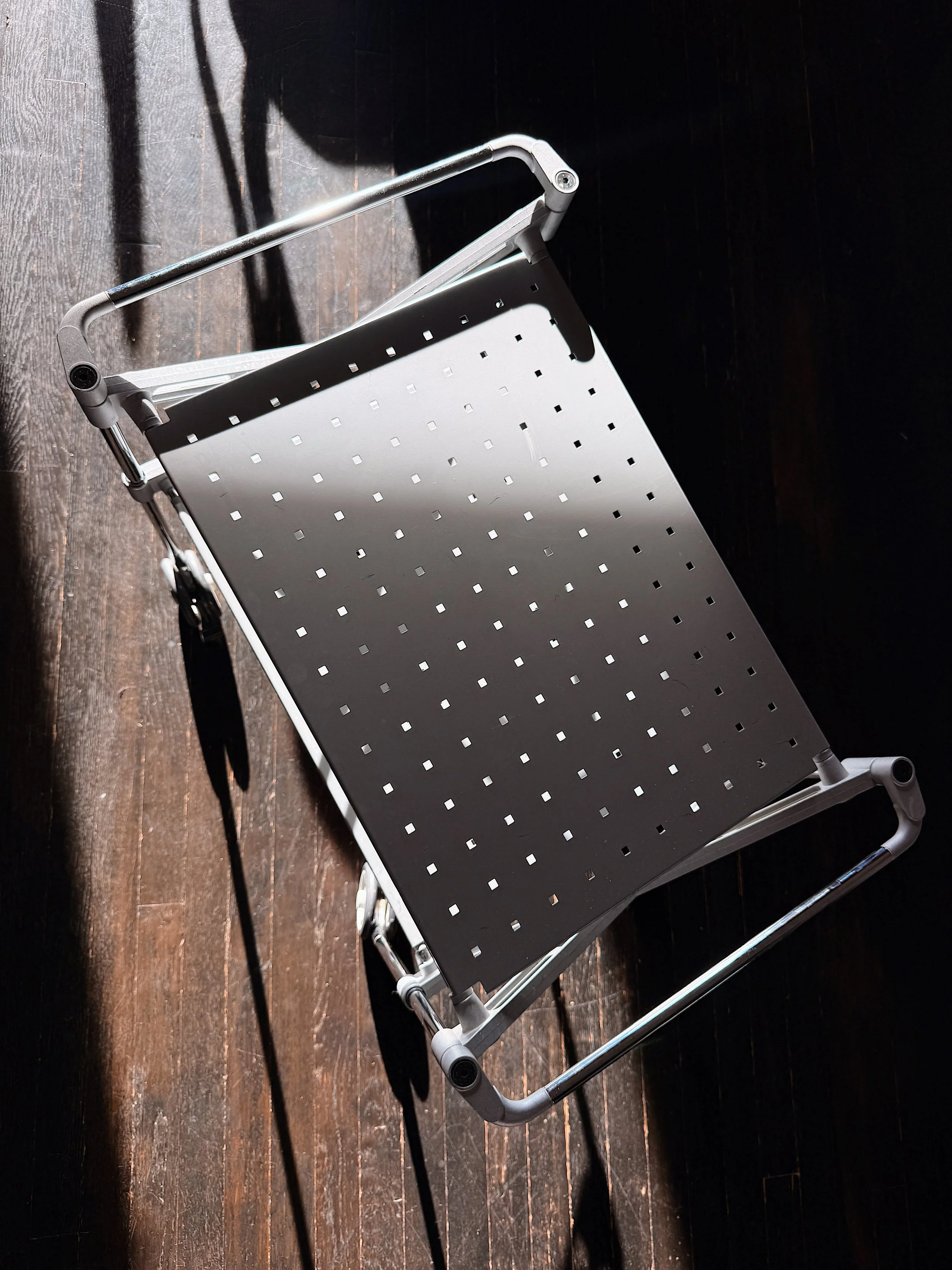 A silver folding platform cart with a perforated black surface, positioned on a wooden floor with a dark wall background.