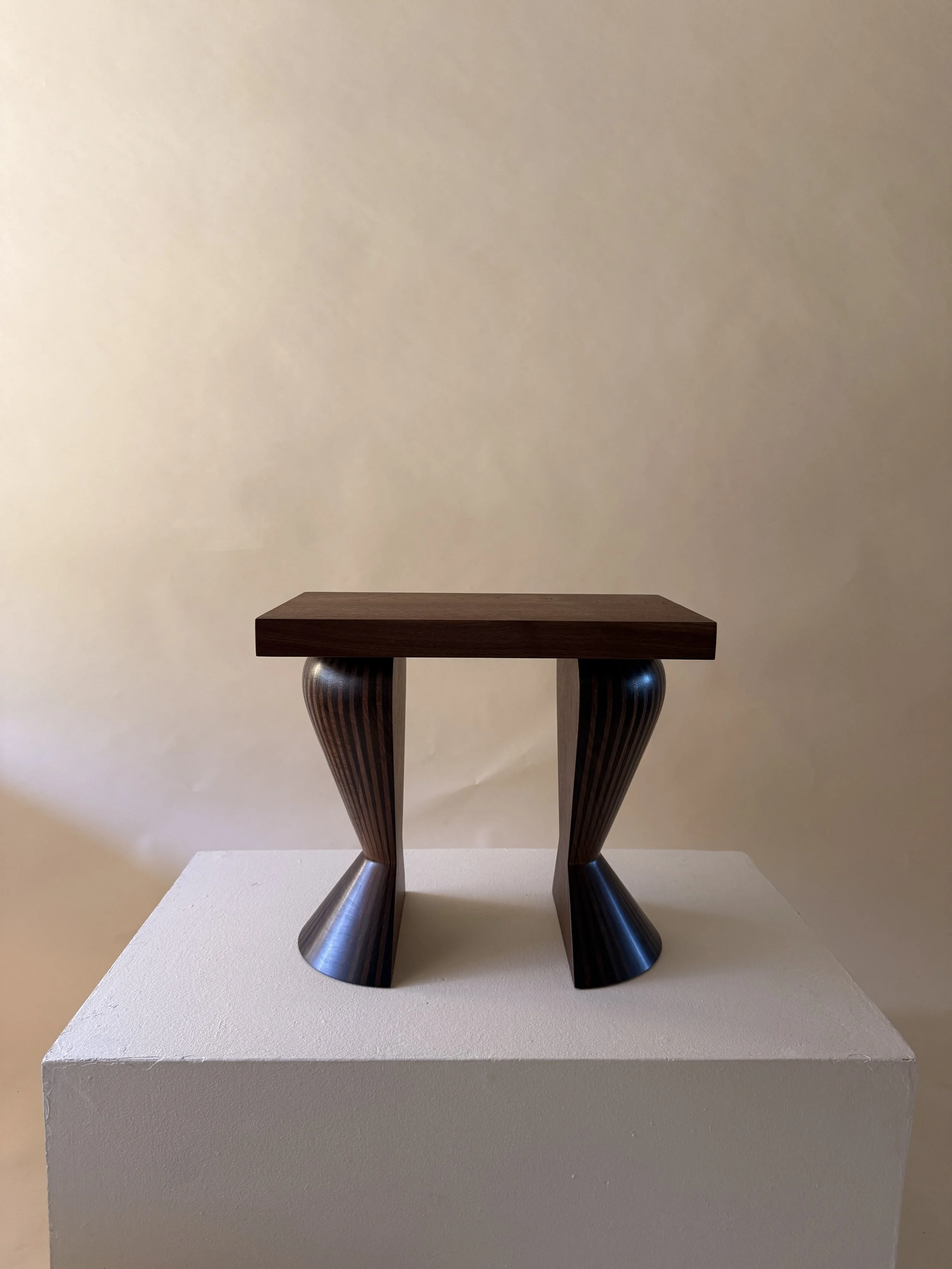 A modern side table with a dark wooden square top and curved wooden legs, supported by metallic bases, placed on a white pedestal against a beige wall.