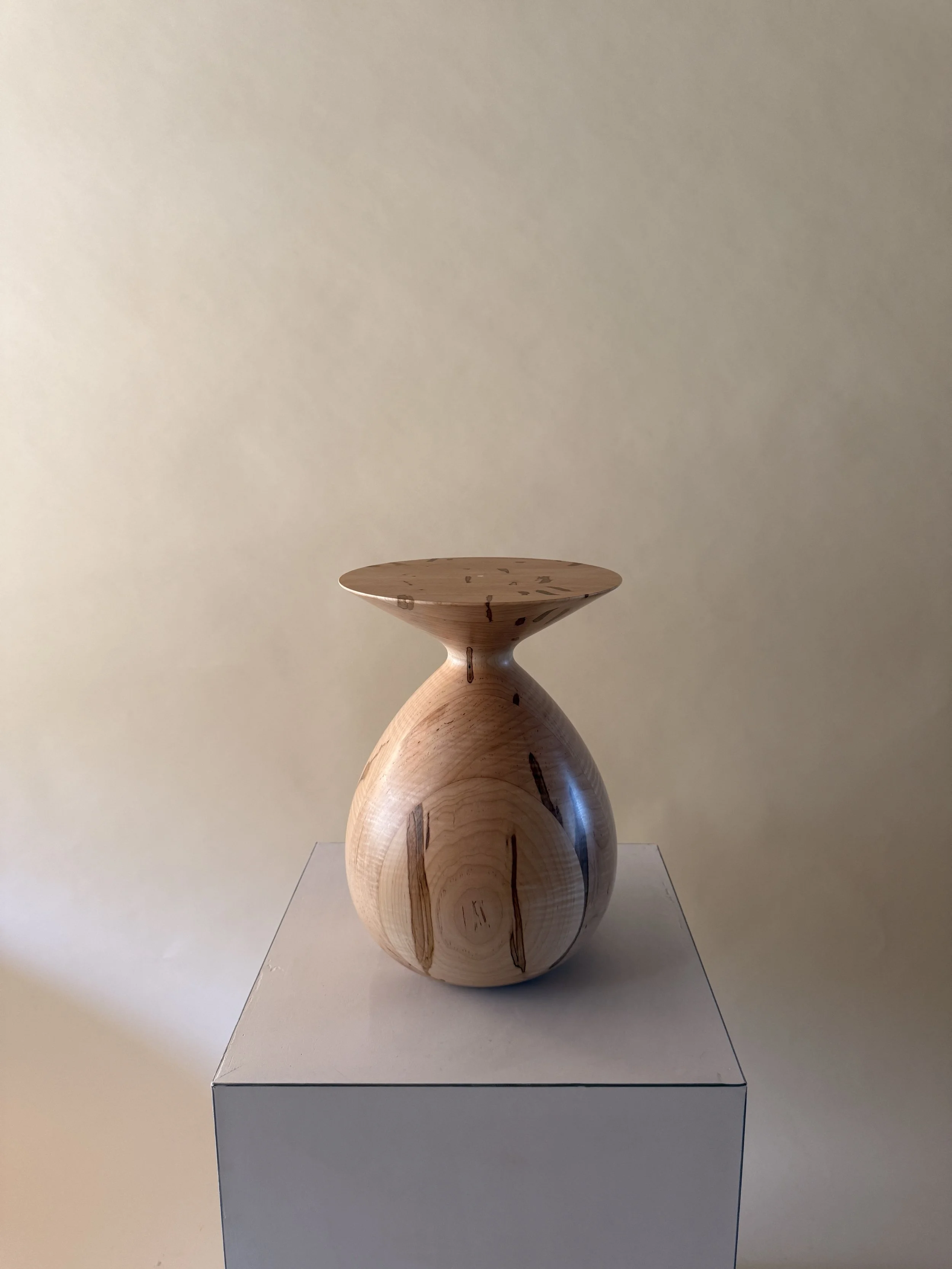 A wooden sculpture consisting of overlapping spherical shapes, with a flat, circular top surface, displayed on a white pedestal against a plain beige wall.