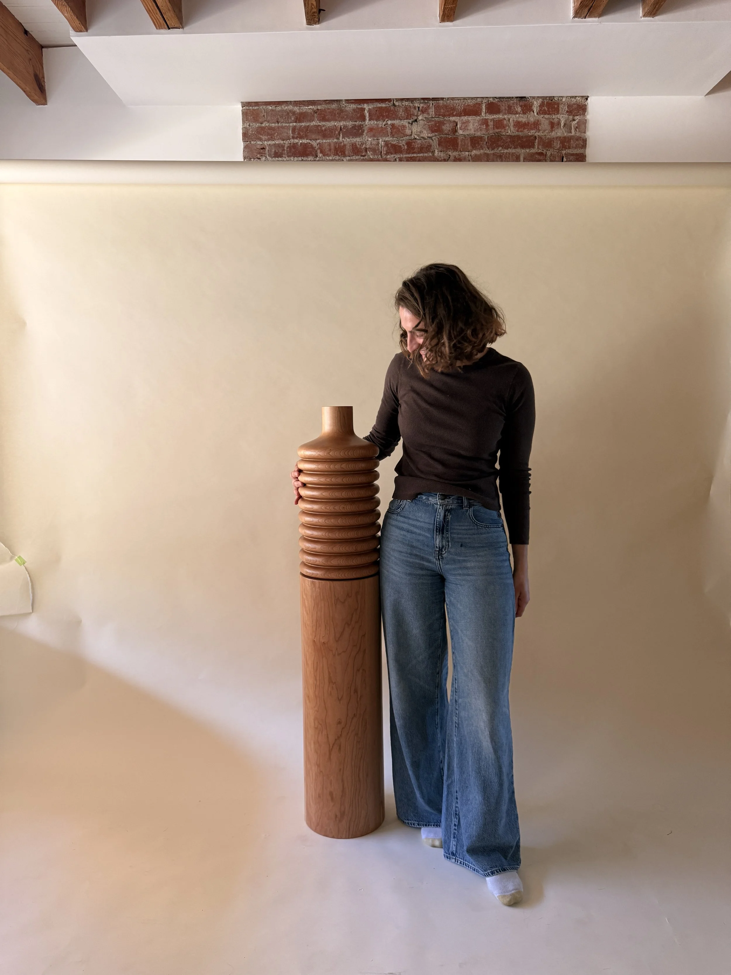 A person with shoulder-length hair wearing a black long-sleeve shirt and wide-leg jeans standing in front of a white wall with exposed brick and wooden ceiling beams, holding a wooden sculpture with stacked, rounded rings.