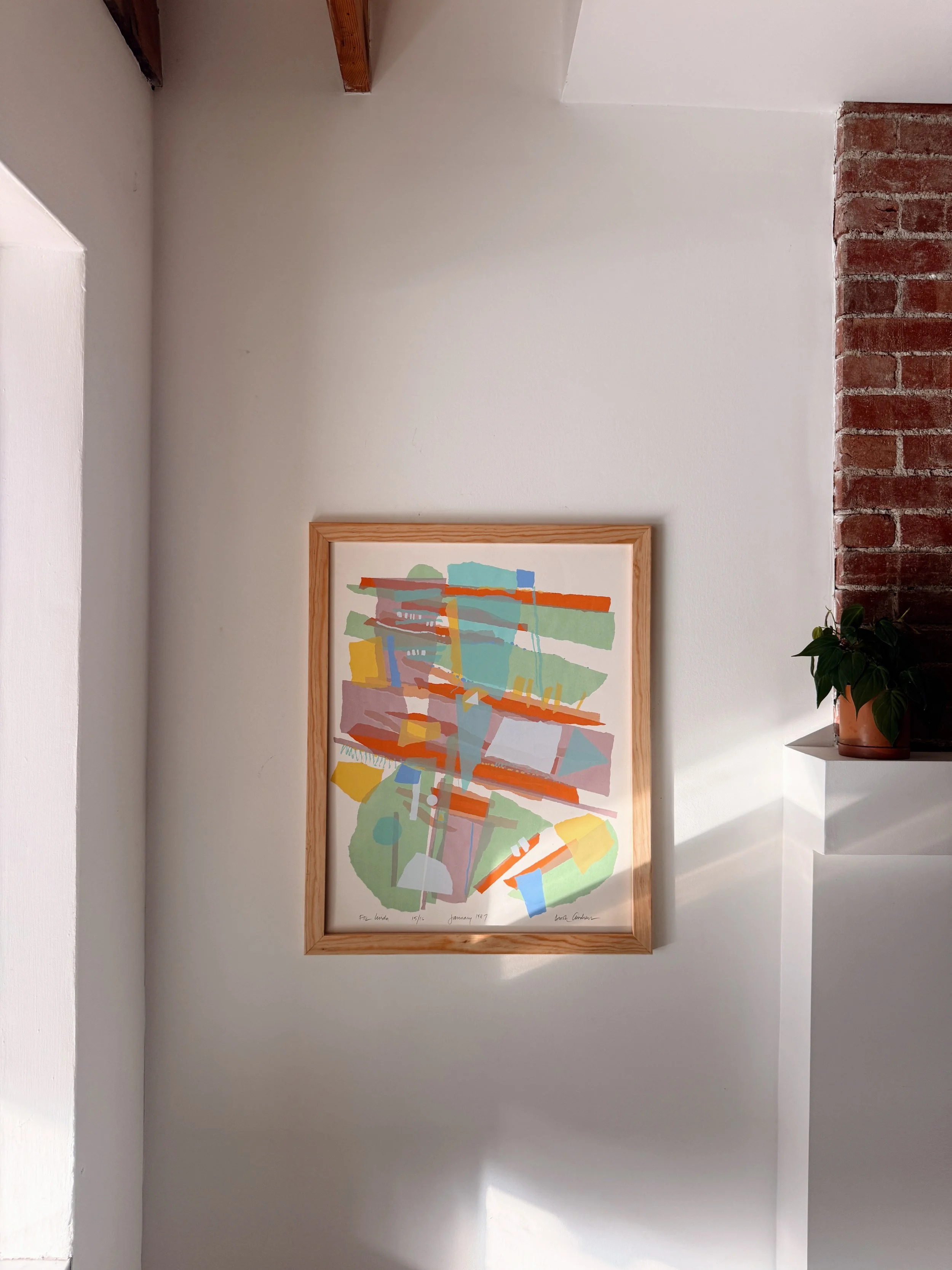An abstract colorful artwork in a wooden frame hanging on a white wall, with a brick column to the right and a potted plant nearby.