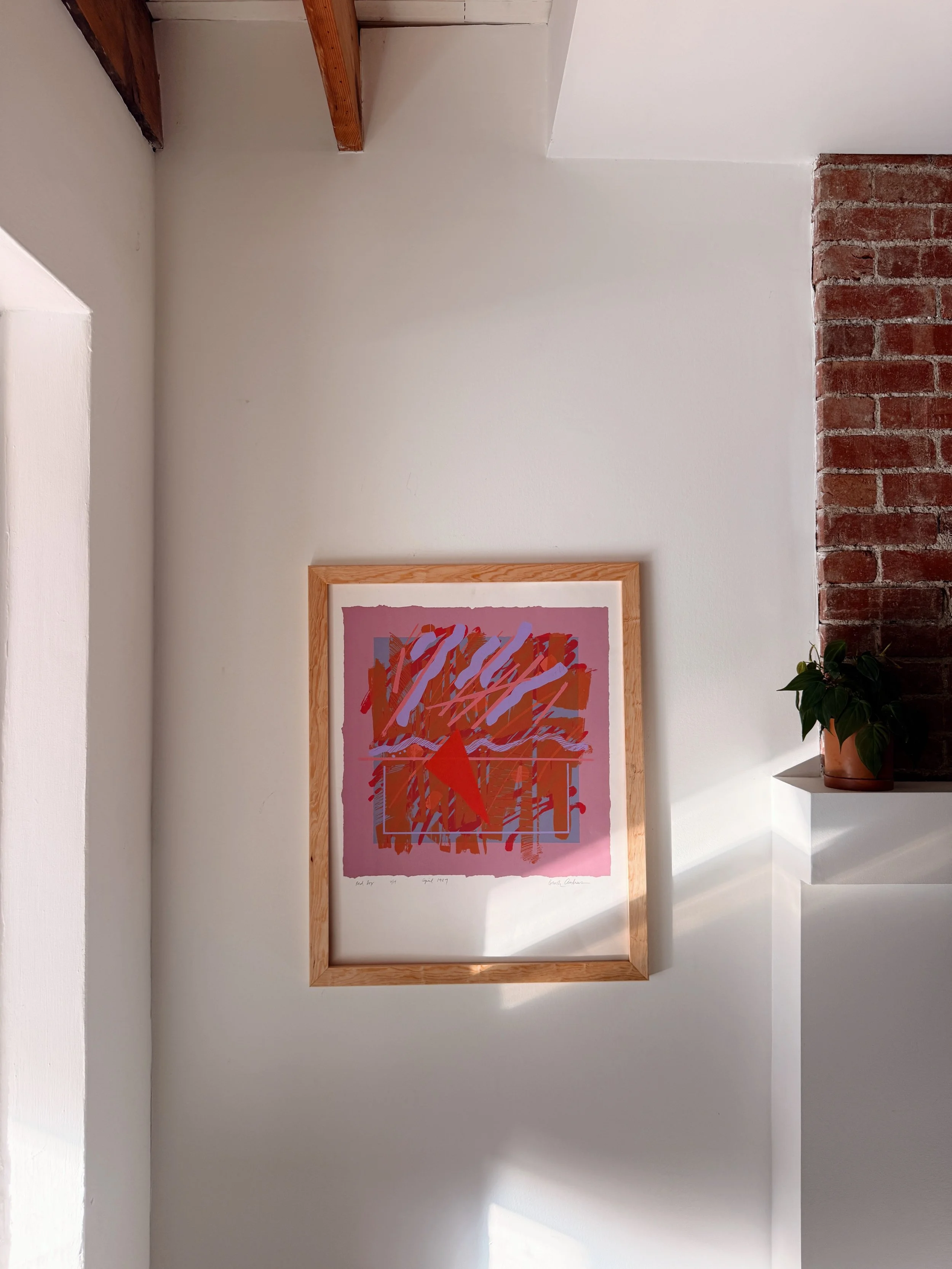 A framed abstract artwork with orange and purple scribbles on a pink background hanging on a white wall near a brick column, with a potted plant on a white shelf to the right.